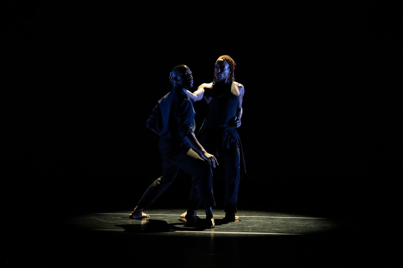 Two dancers performing a duet inside a pool of light on a dark stage, one reaching out to touch the other's head.