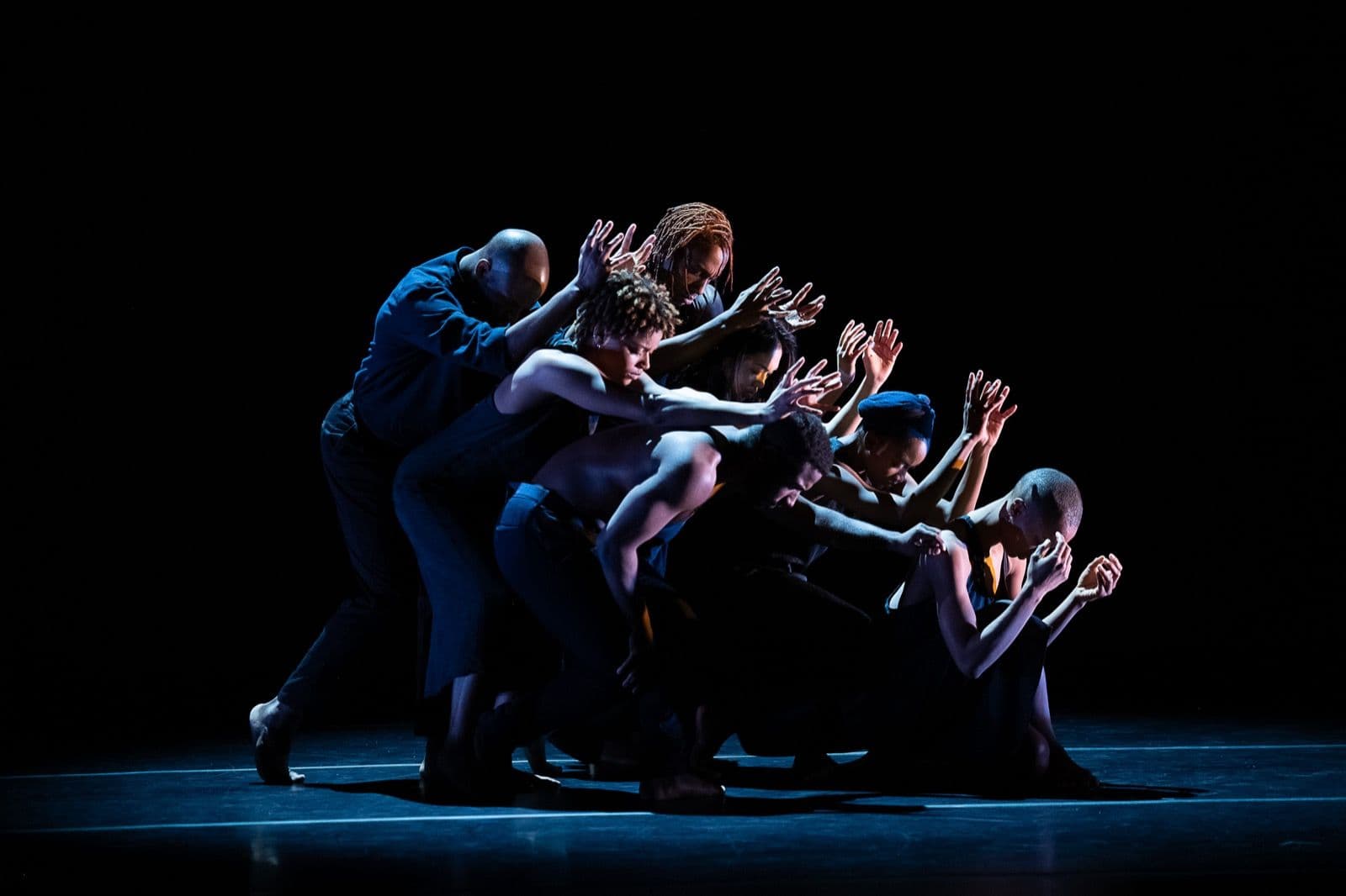 A group of dancers leaning forward in a compact, diagonal formation with arms extended outward under dramatic lighting.