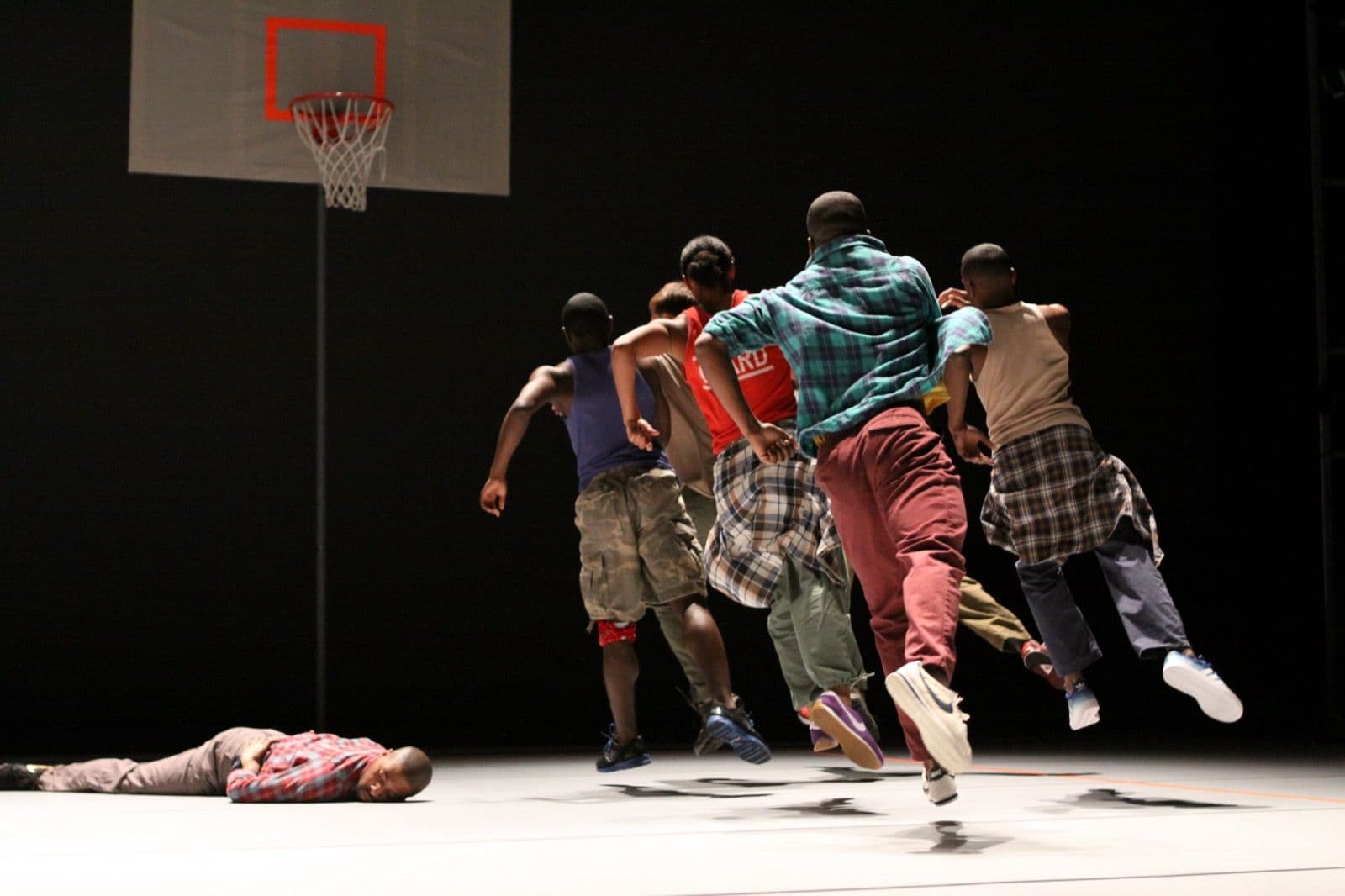 Dancers in mid-air jumping toward a basketball hoop while one dancer lies face-down on the stage floor.