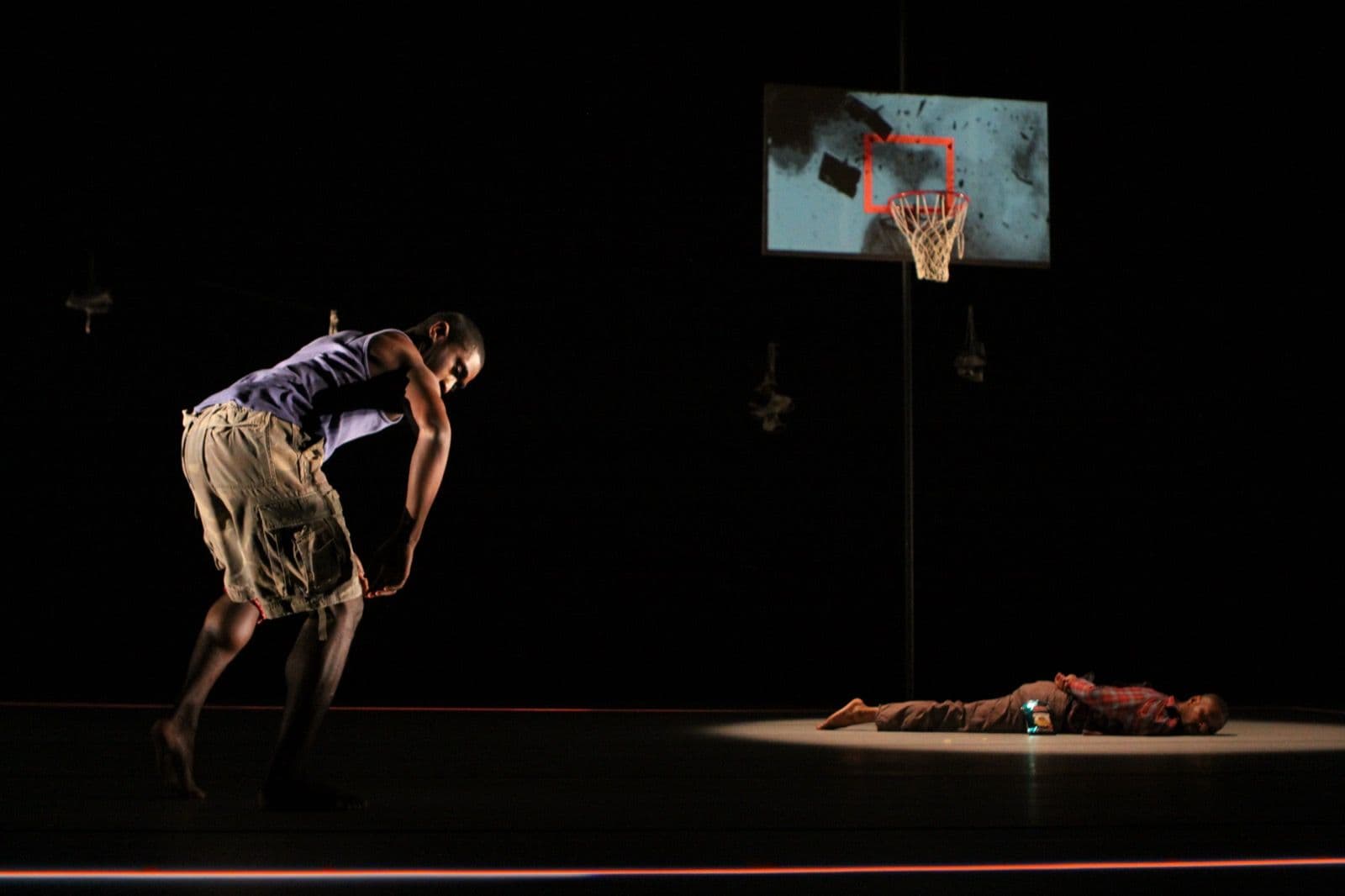 A dancer leaning forward on a dark stage near a basketball hoop while another lies prone under a spotlight.