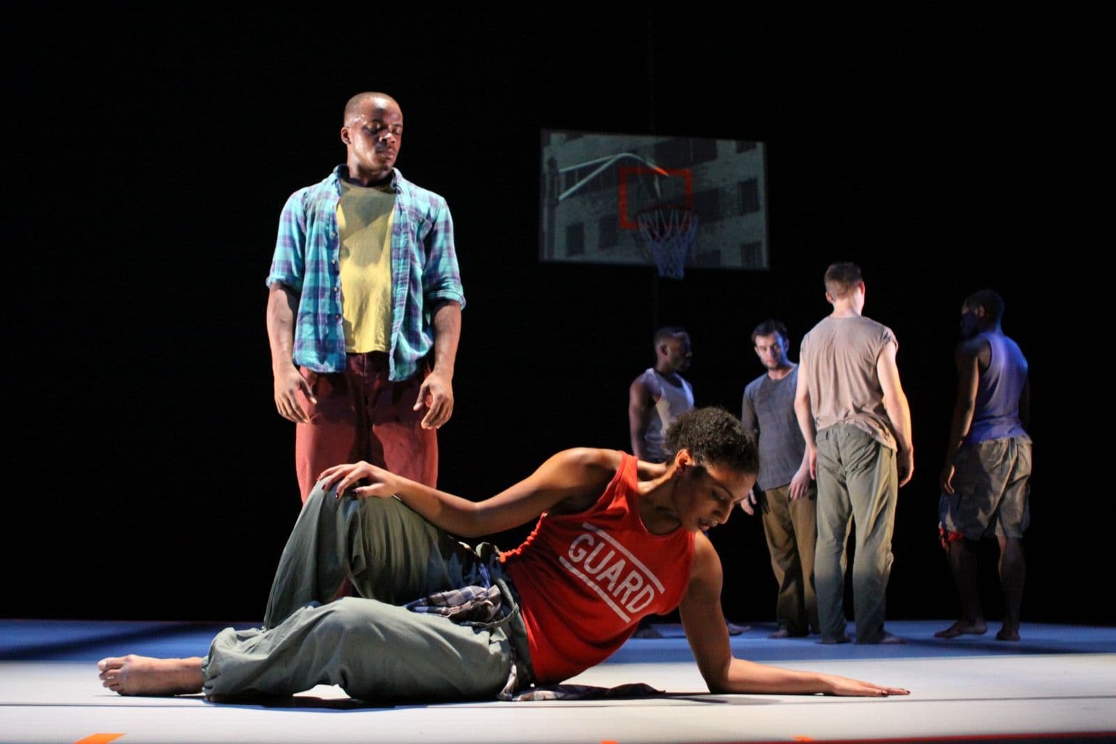 Dancers on stage under dark lighting, with one woman lying on the floor in the foreground and a man in a plaid shirt standing nearby.