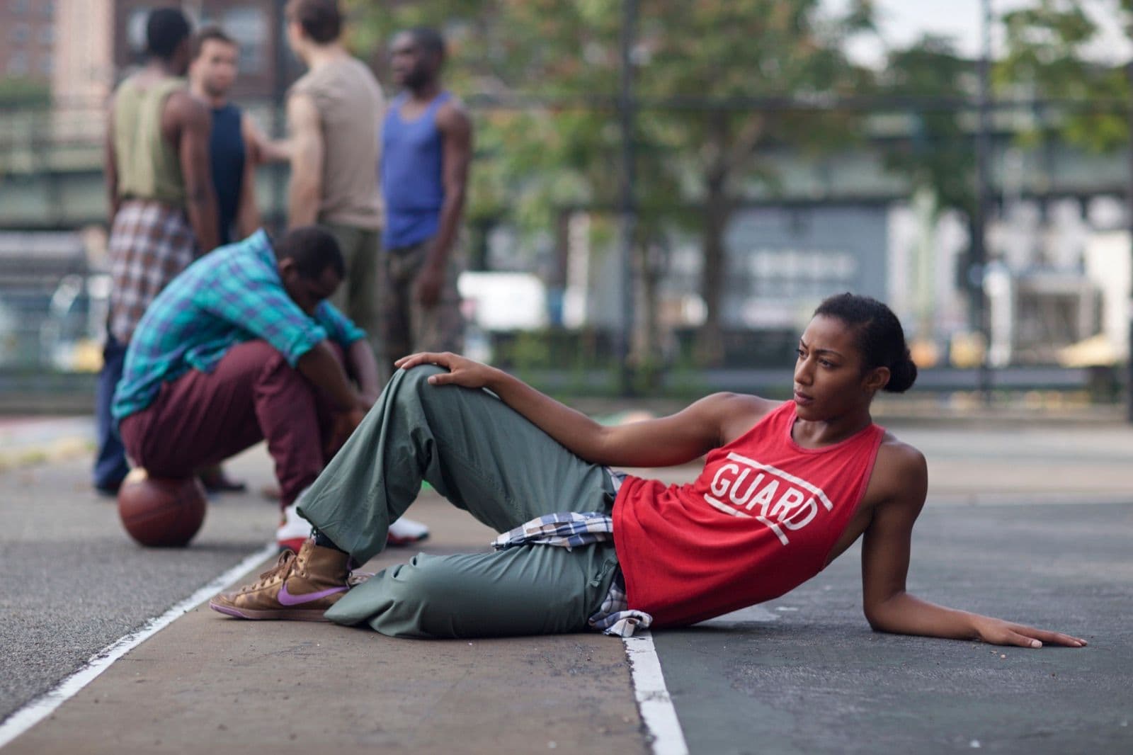 A dancer reclining on an outdoor basketball court while other dancers gather in the background.