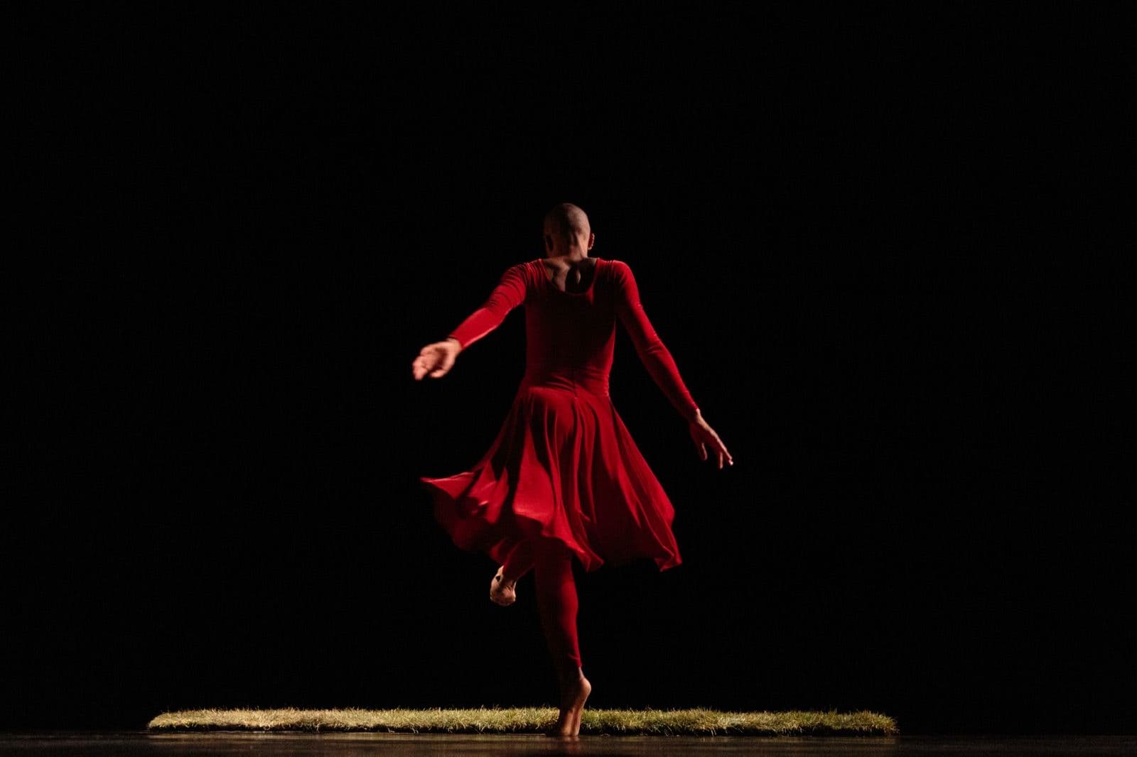 Dancer performing in a long-sleeved red dress from behind, captured mid-spin on a strip of grass set against a dark background.