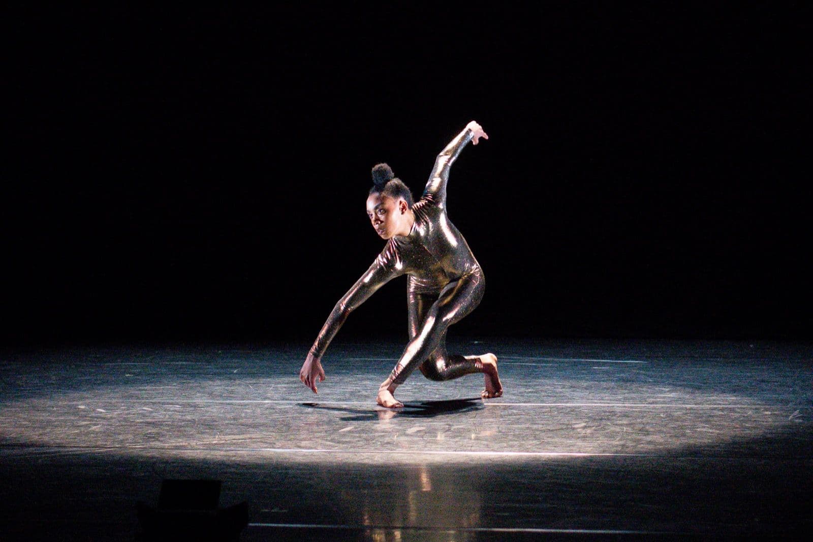 Dancer Marcella Lewis crouching low to the stage floor in a pool of light during a solo performance.