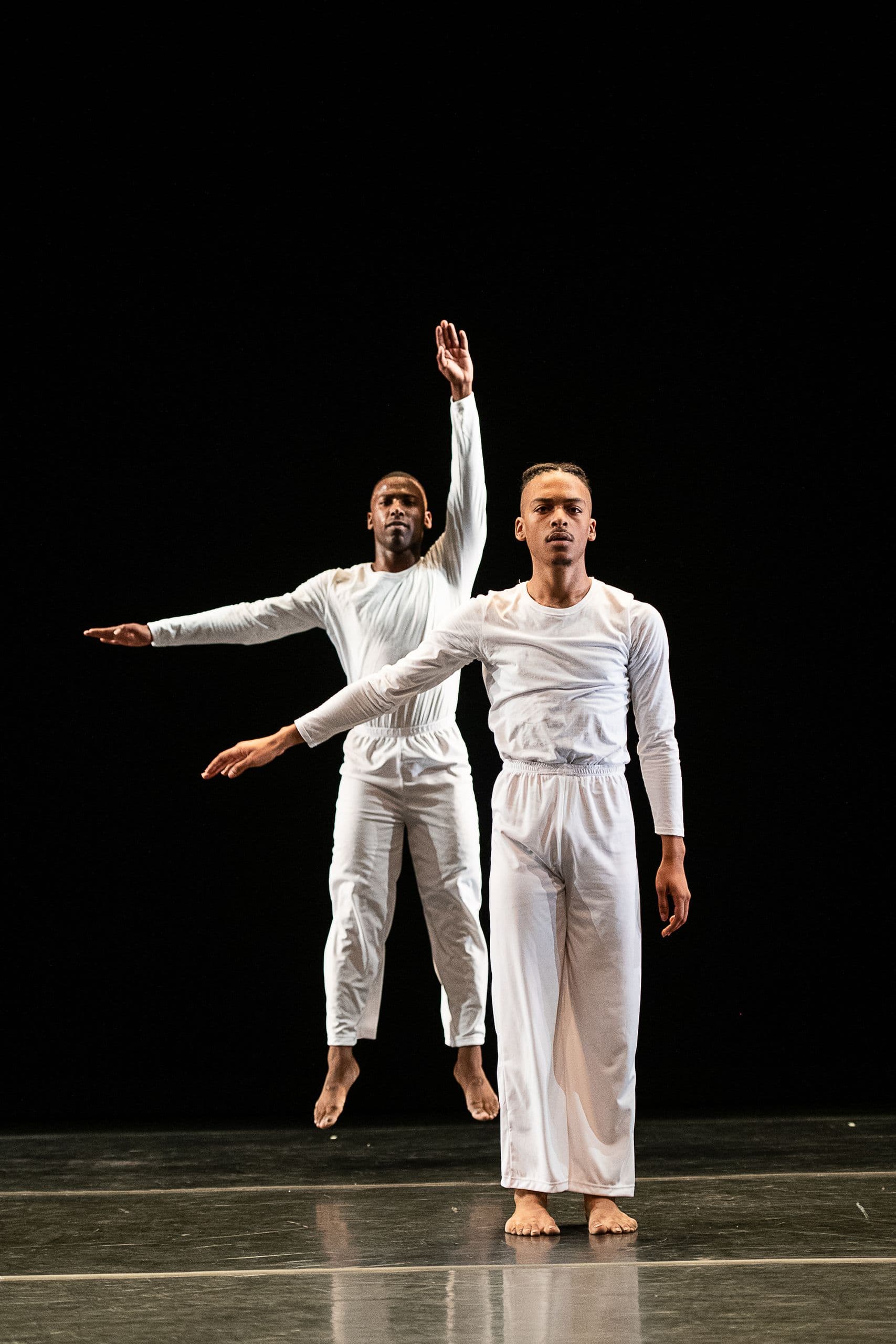 Two dancers in white stand together on a dark stage, one leaping with arm raised overhead behind a second dancer standing still with arm extended sideways, from 'Solo Olos.