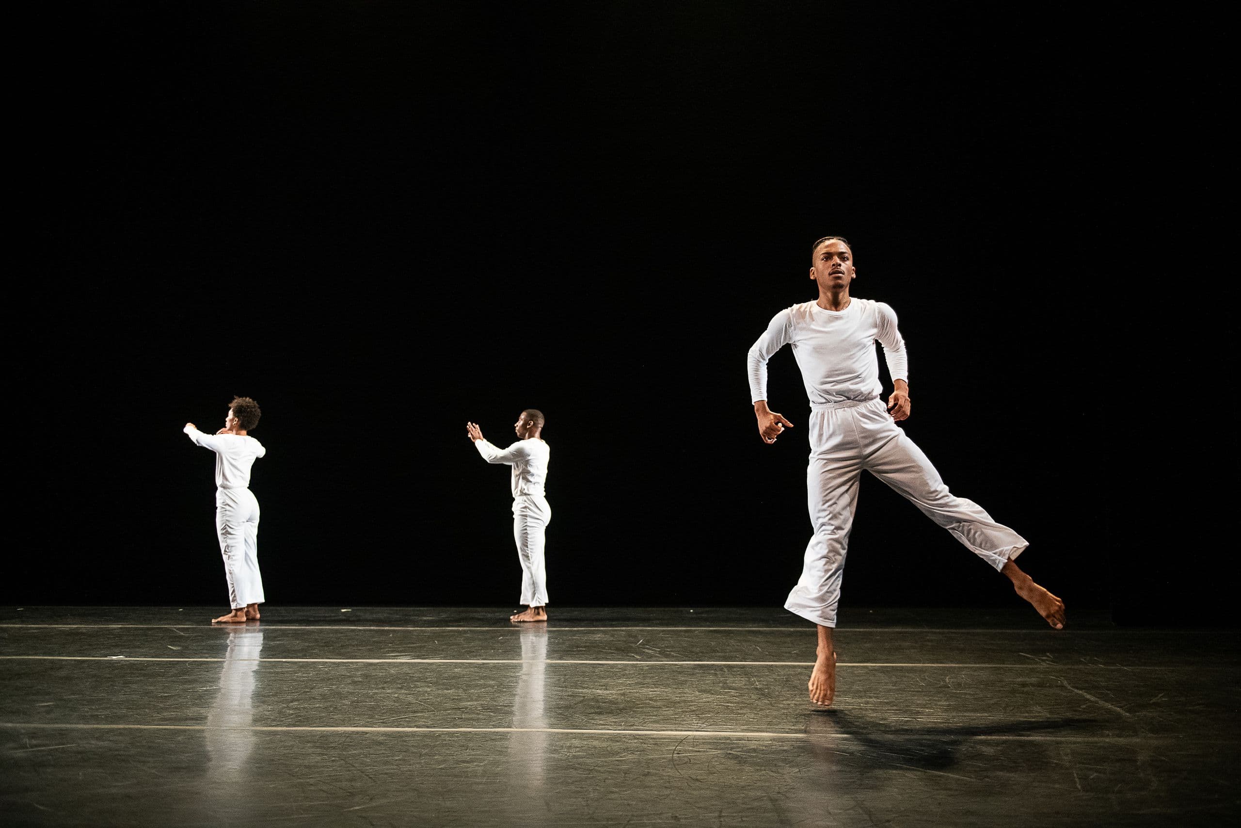 Three dancers in white step forward across a dark stage, the closest dancer mid-stride with one leg extended behind him, from 'Solo Olos.'
