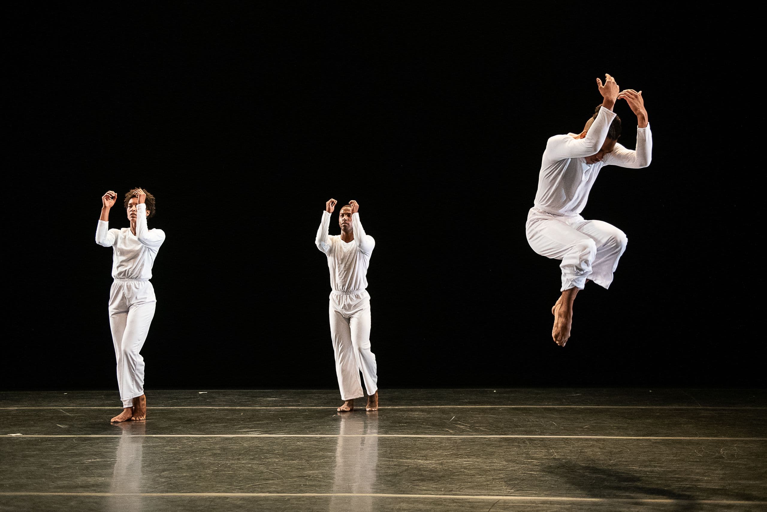 Three dancers in white on a dark stage: two stand with arms raised, while a third leaps mid-air with arms crossed overhead, from 'Solo Olos'.
