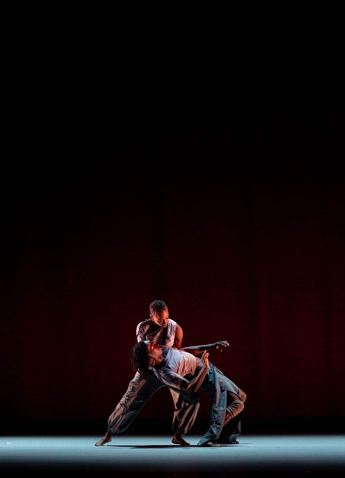 Two dancers from A.I.M by Kyle Abraham perform a dramatic, leaning duet under a single overhead spotlight on a dark stage.