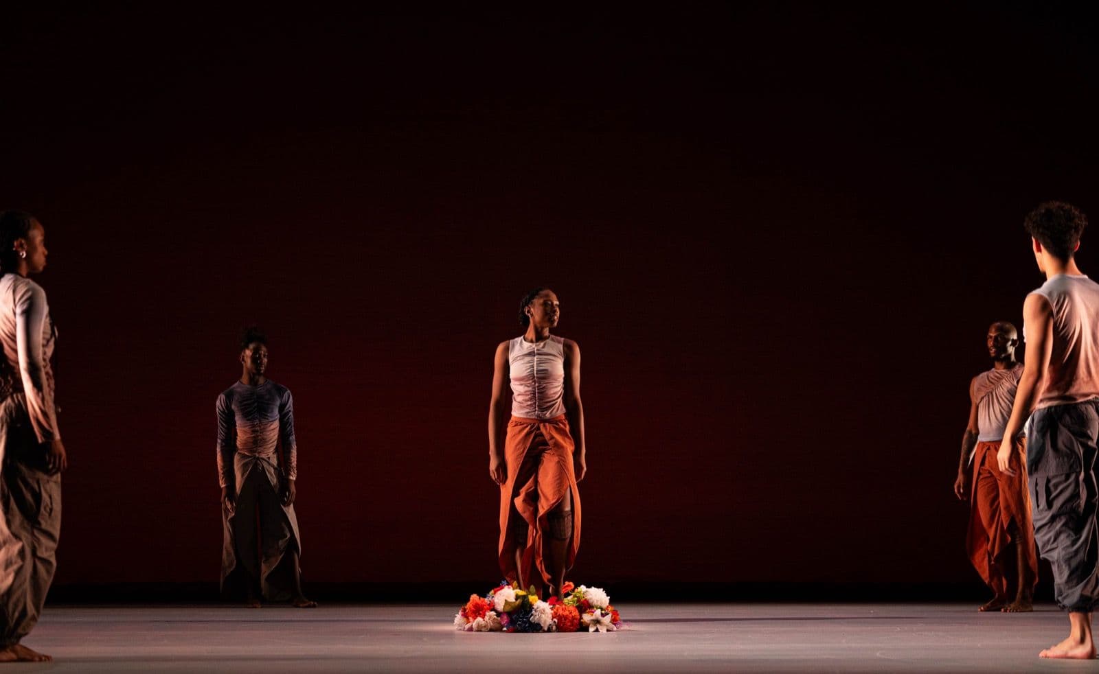 A dancer stands centered over a pile of colorful flowers on stage, surrounded by ensemble members standing in a wide semicircle.