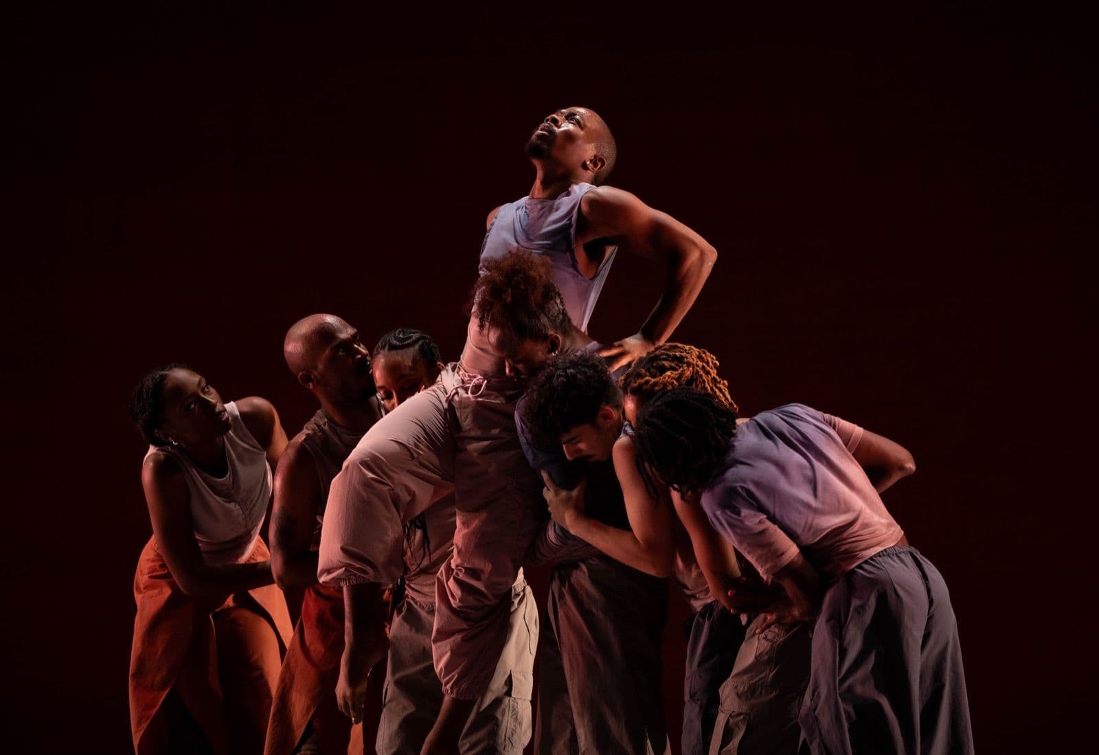 A group of dancers closely surrounds and lifts a central dancer reaching toward the ceiling under warm stage lighting.