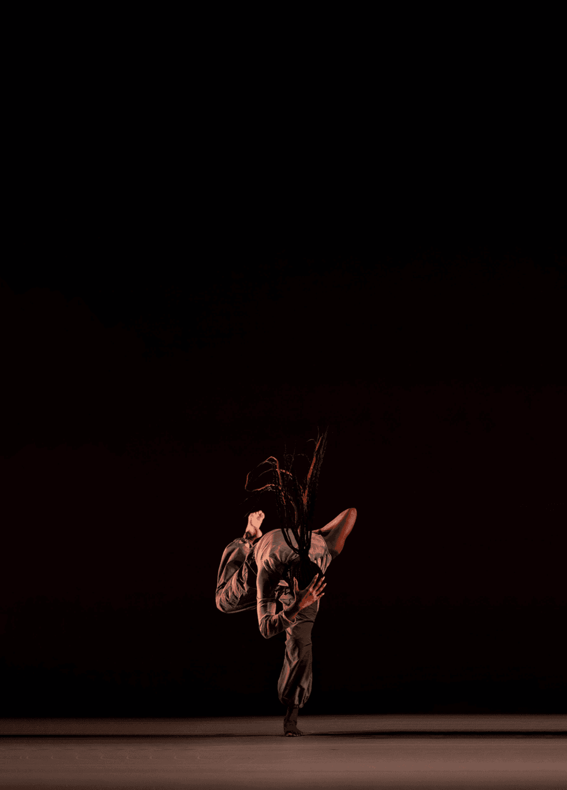A solo dancer performs a dynamic, inverted movement on a dark stage, whipping braided hair forward mid-motion.