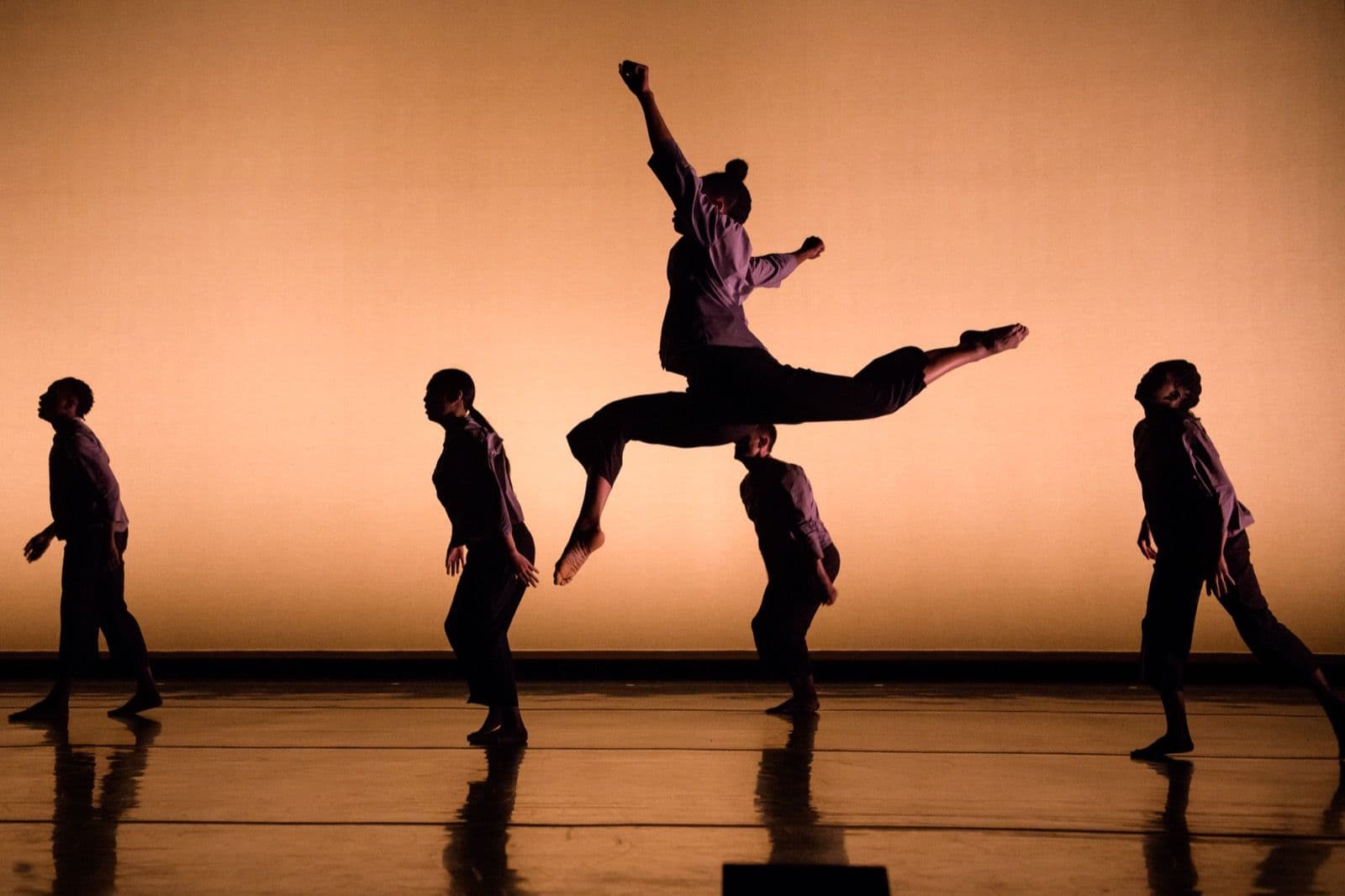 Silhouetted dance ensemble on stage with a back-lit warm orange background as one dancer leaps dynamically in mid-air.