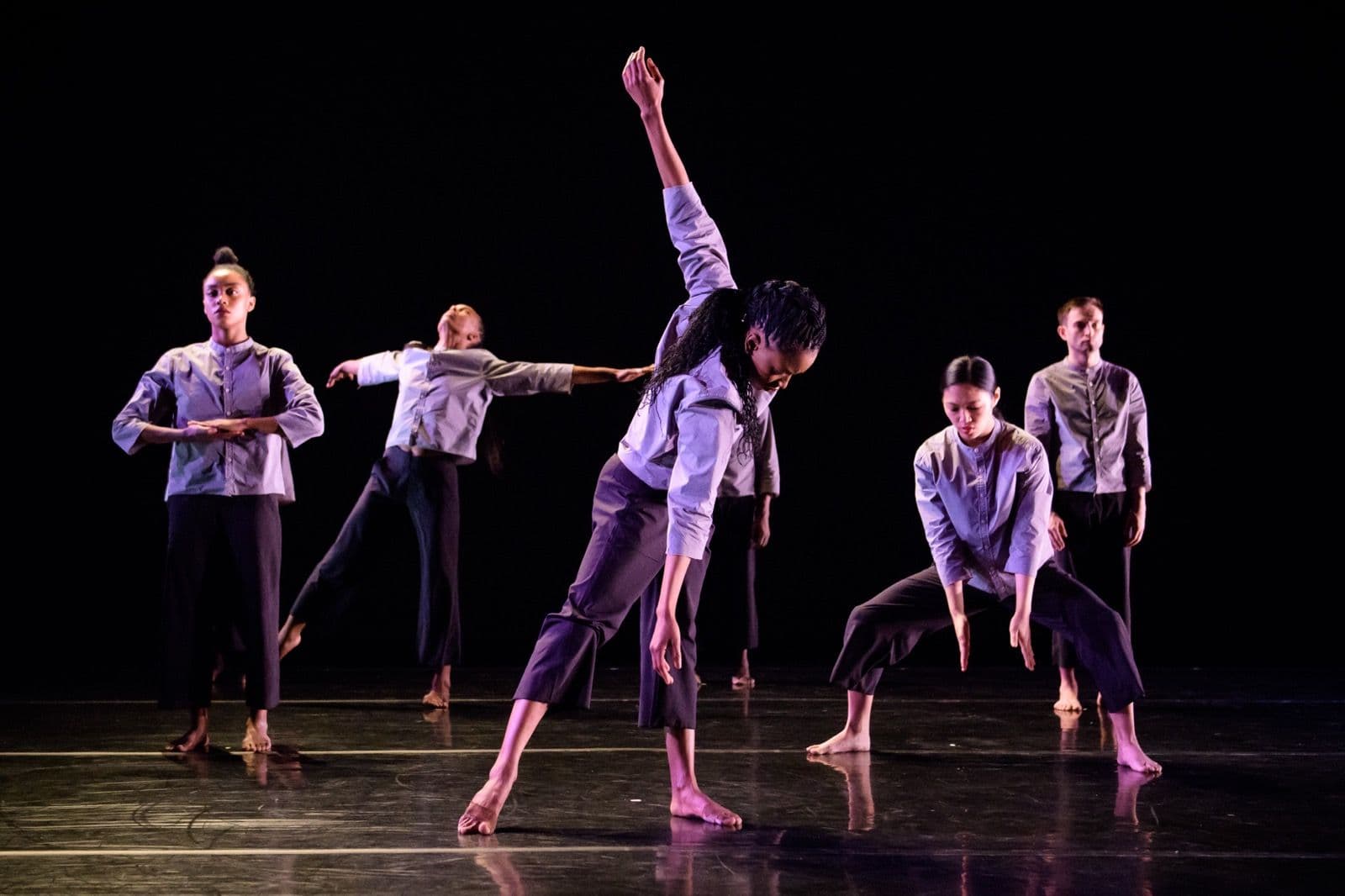 Five contemporary dancers in light shirts and dark trousers performing grounded, angular poses under dramatic stage lighting.