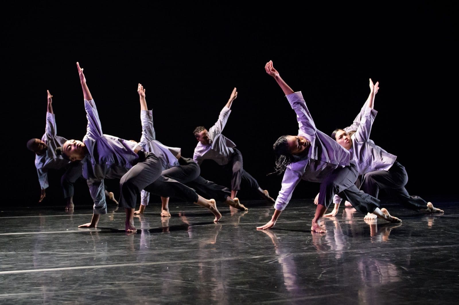 Dance ensemble leaning low to the side with outstretched arms against a dark backdrop.
