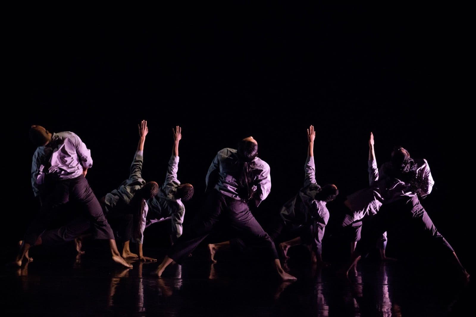 Dancers viewed from behind in a dramatic stage lights, arching backward with arms raised toward the ceiling.