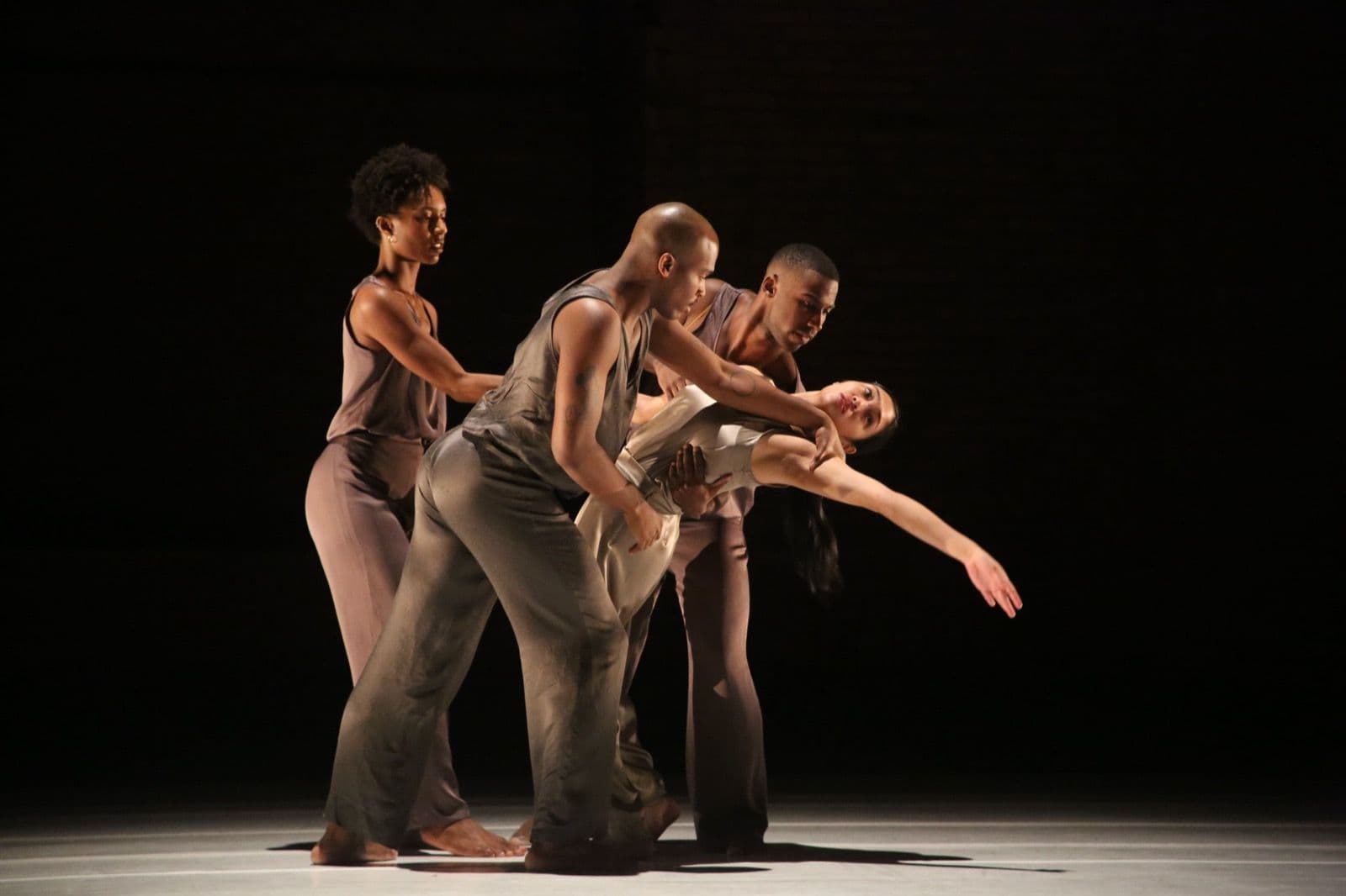 Four contemporary dancers in neutral-toned outfits perform a synchronized, expressive movement on a dimly lit stage.