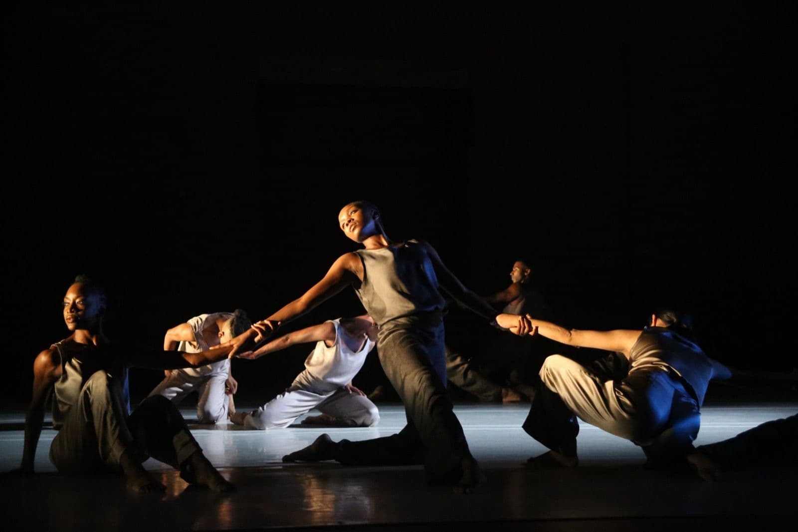 A group of dancers in grey and white attire perform an interconnected, dramatic pose across a lit stage against a dark background.