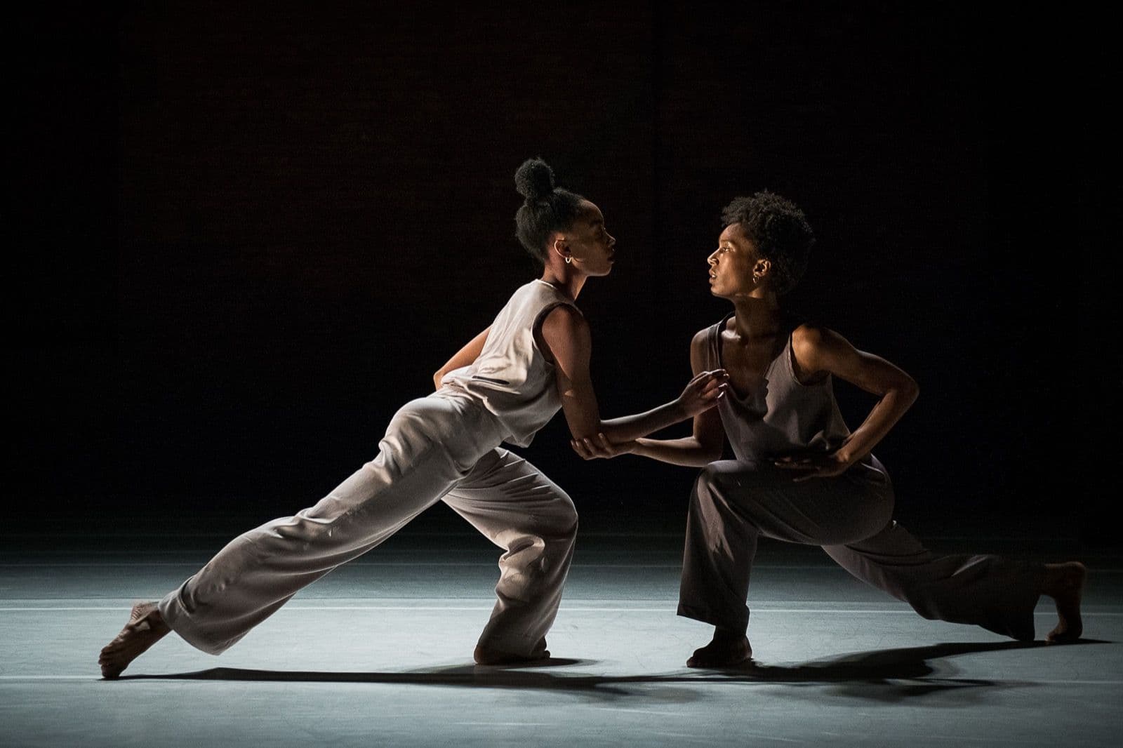 Two female dancers face each other in low, lunged poses during an intimate contemporary dance duet under dramatic stage lighting.