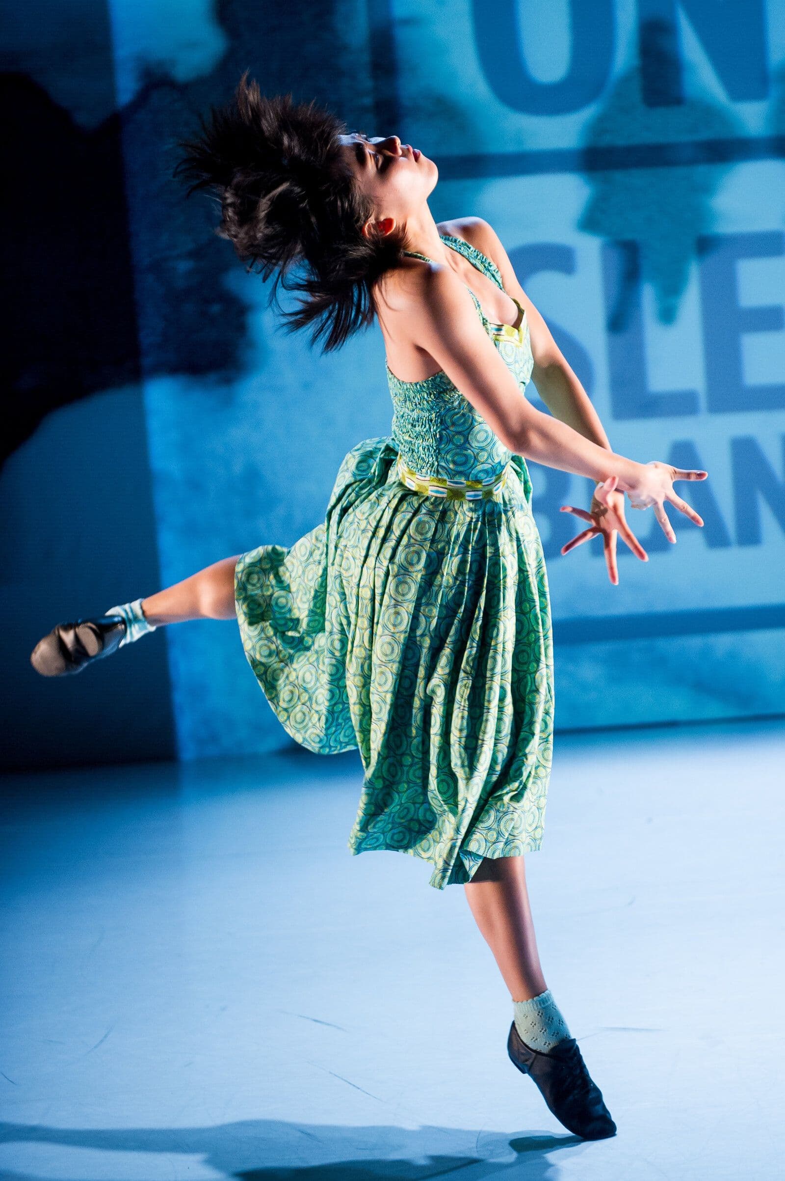Dancer leaps backward with hair flying and one leg extended, wearing a green patterned dress, lit in cool blue tones against a projected backdrop reading "White Area."