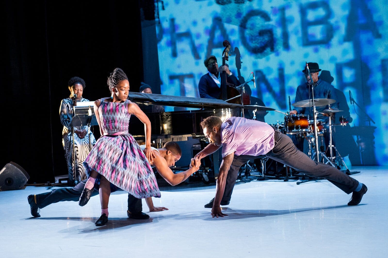 Three dancers in vintage-style patterned clothing move together near a grand piano and live band, one in a full skirt reaching toward two partners in a low lunge, lit in blue stage light with bold projected text overhead.