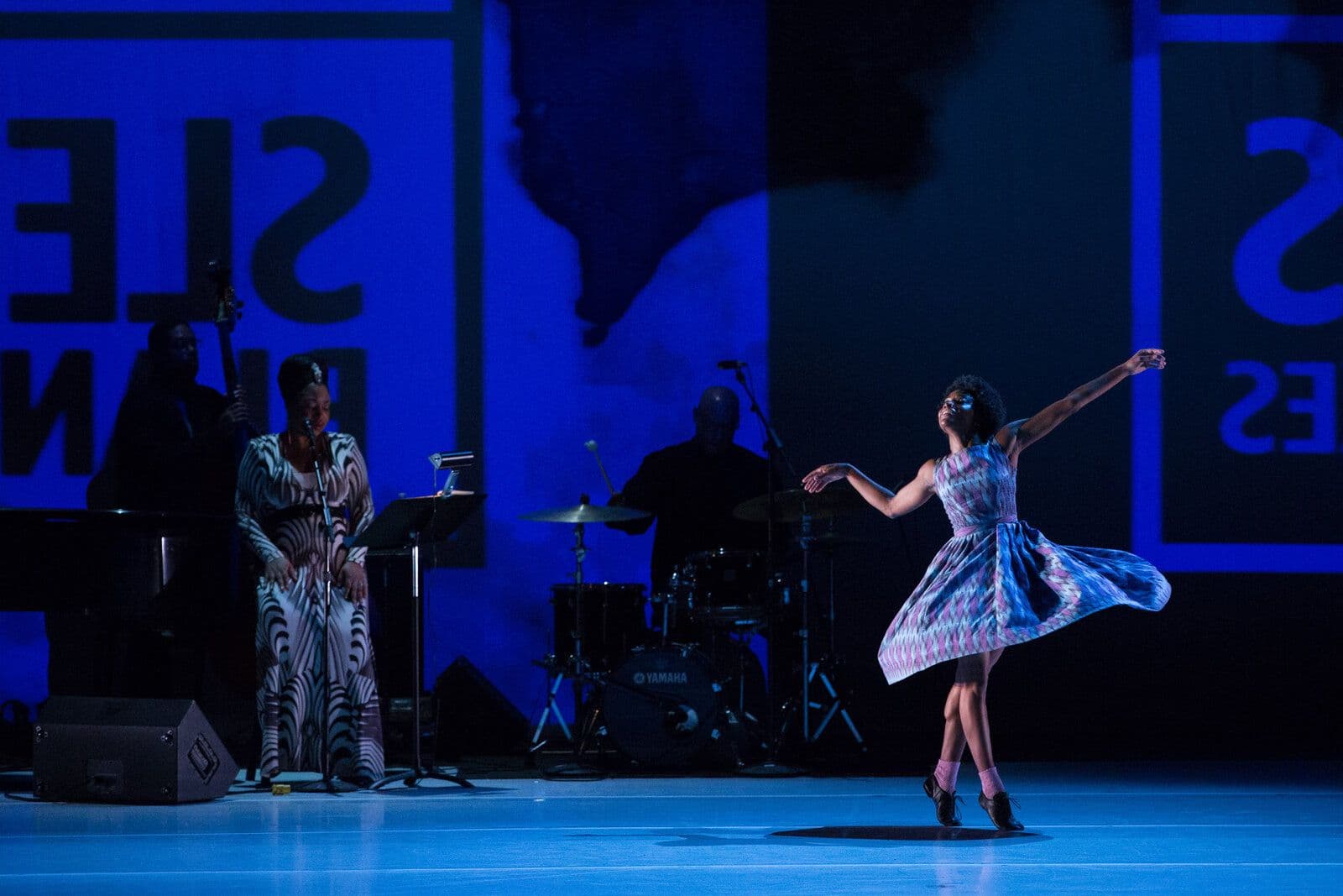 Dancer spins in a swirling patterned dress beside a live band, including a vocalist and drummer, lit in deep blue with large projected lettering in the background.