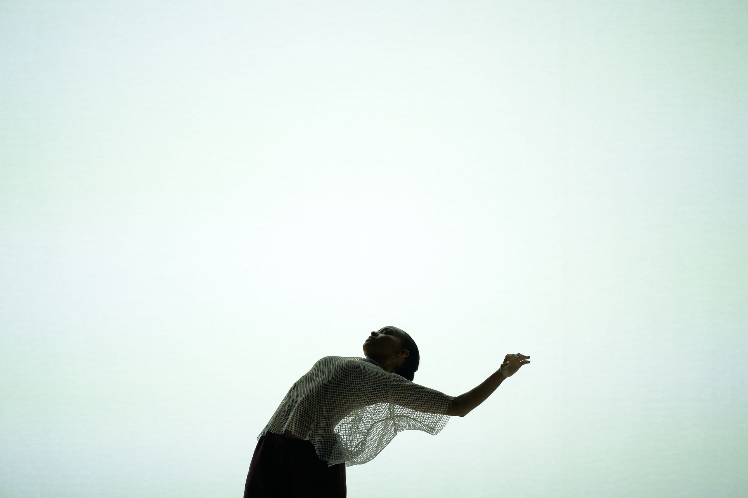 Alysia Johnson reaches their arms above and behind as they arch their back in front of a white skrim.