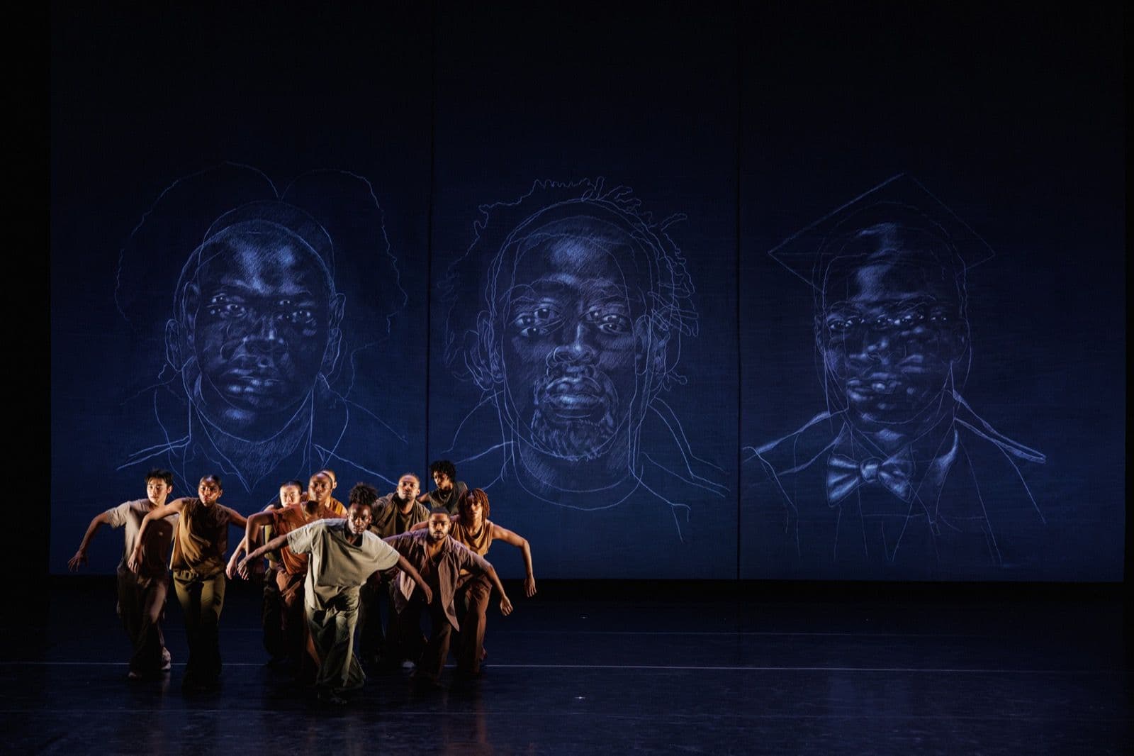 A dance ensemble moves across a dark stage in front of a backdrop featuring three large line-art portrait sketches.