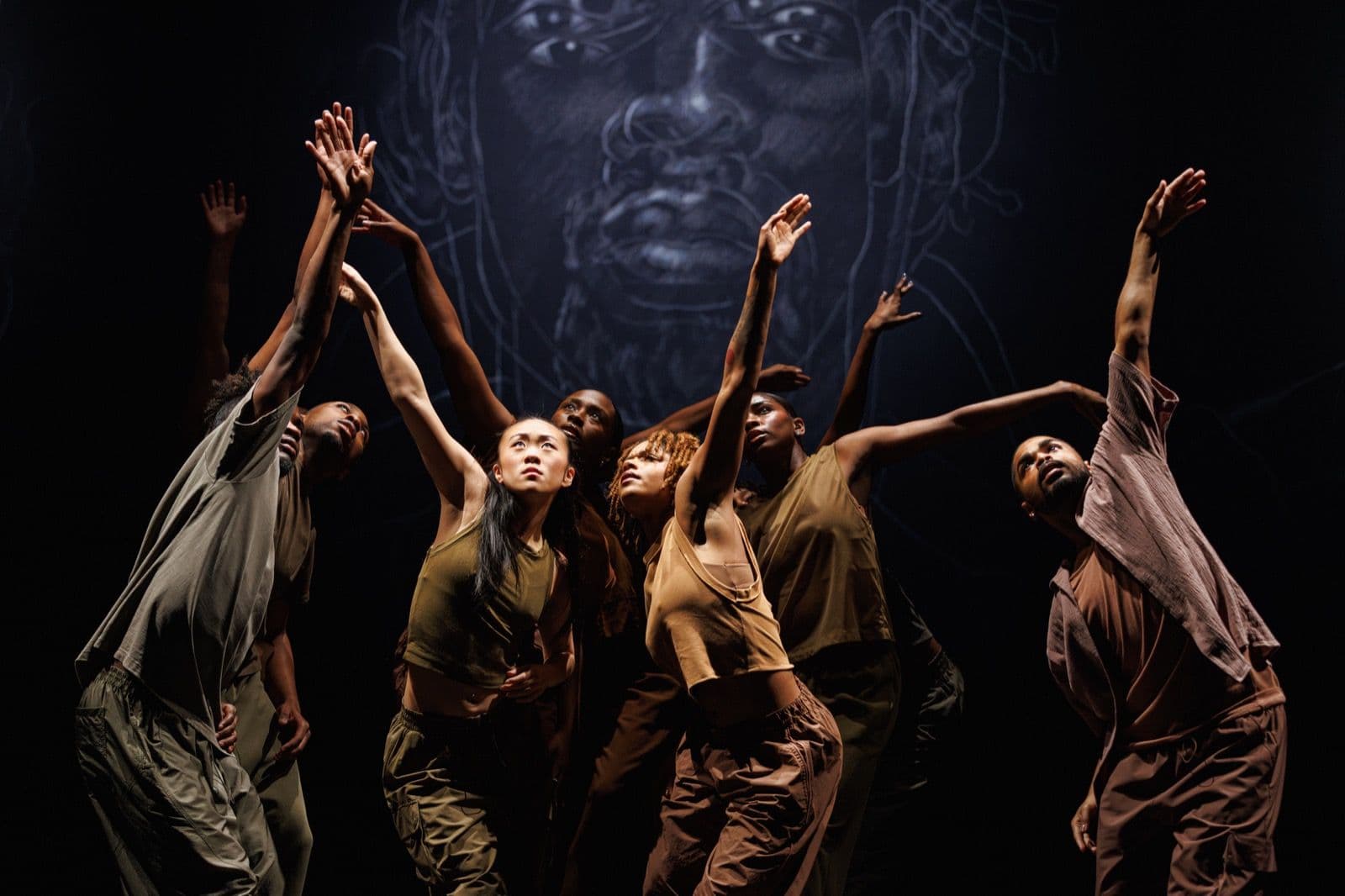 A group of dancers reach upward in unison against a dark backdrop illuminated by a faint portrait design.