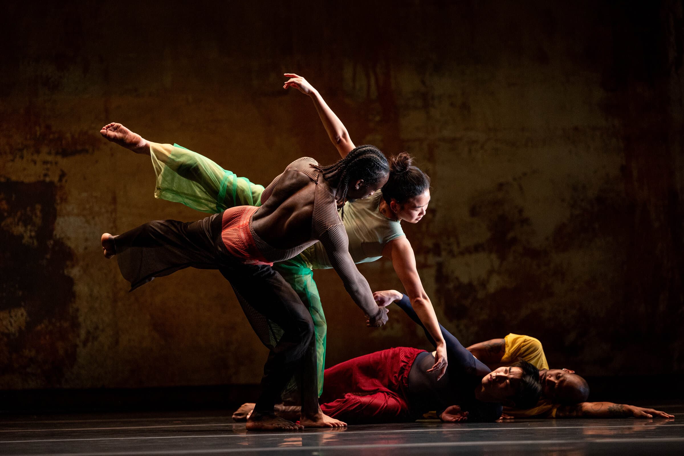 Four dancers in colorful mesh and fabric costumes caught mid-fall around a dancer lying on the floor, dramatic rust-toned lighting.