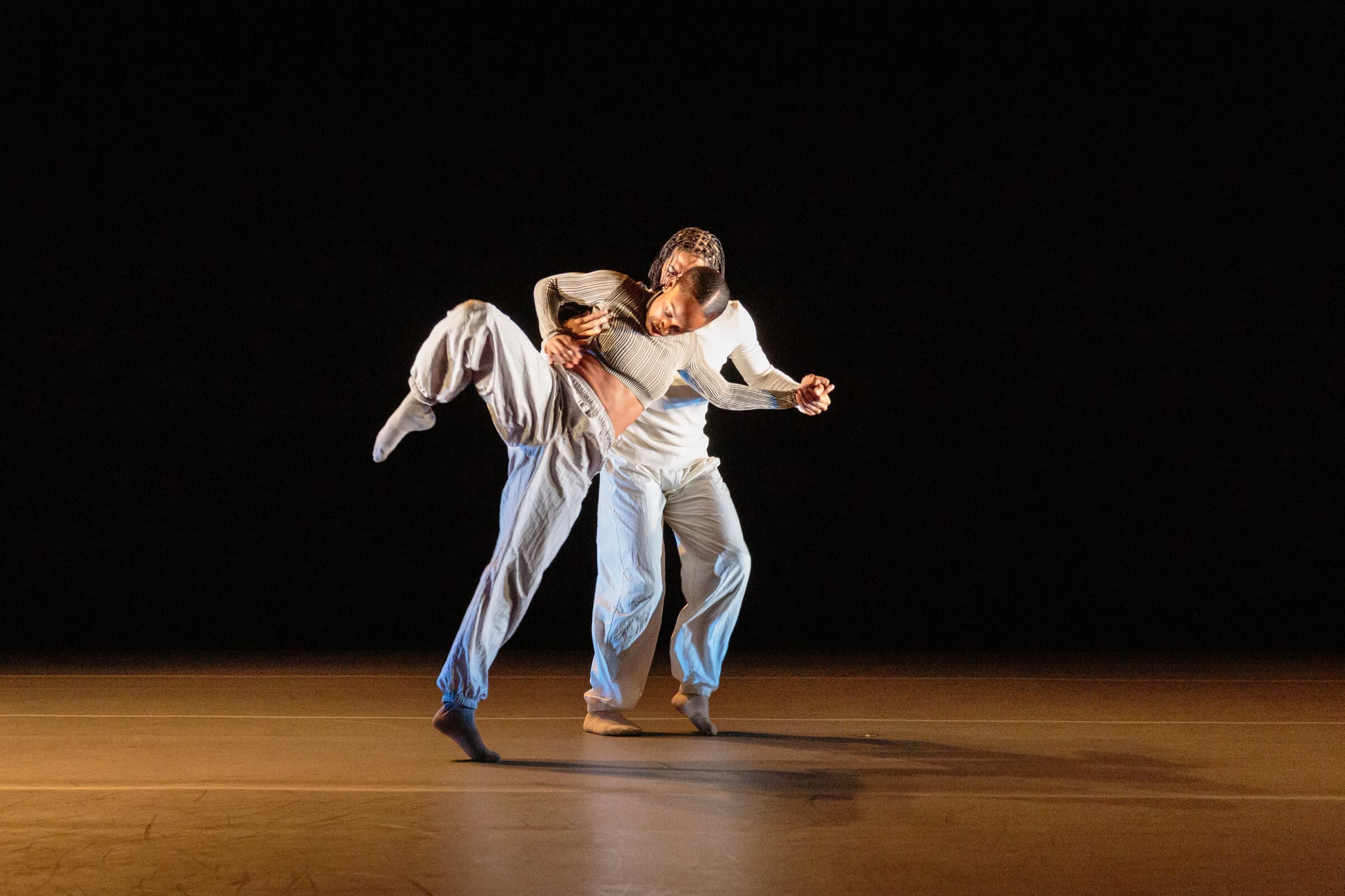Two dancers, Karmel Small and Keturah Stephen, in cream clothing, one lifting a leg high while embracing the other on a dark stage