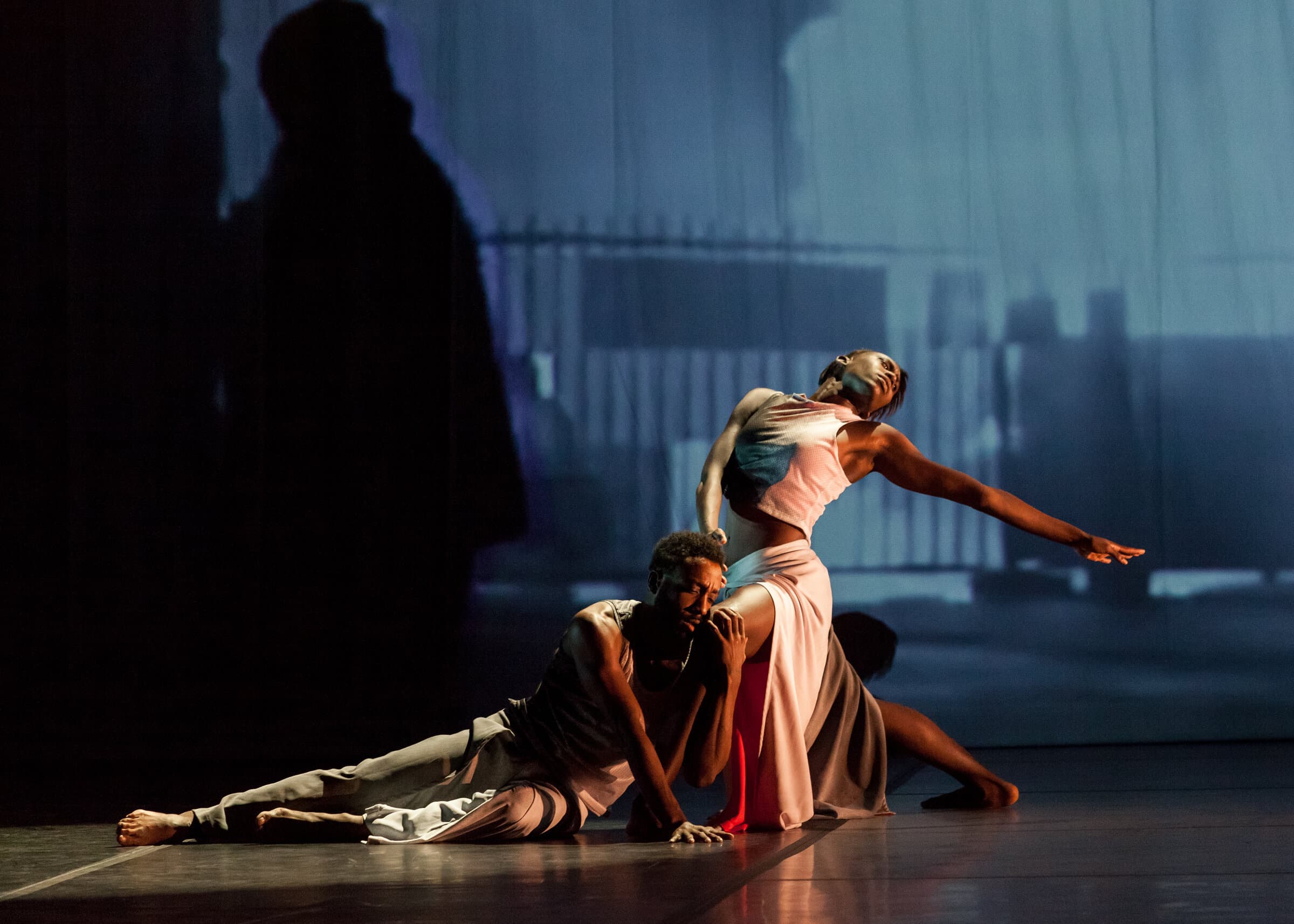 Dancers Jae Neal and Penda N'diaye, one lifted in an extended backbend with arm outstretched, another crouched beside her, blue-lit stage with fence-like backdrop projection.