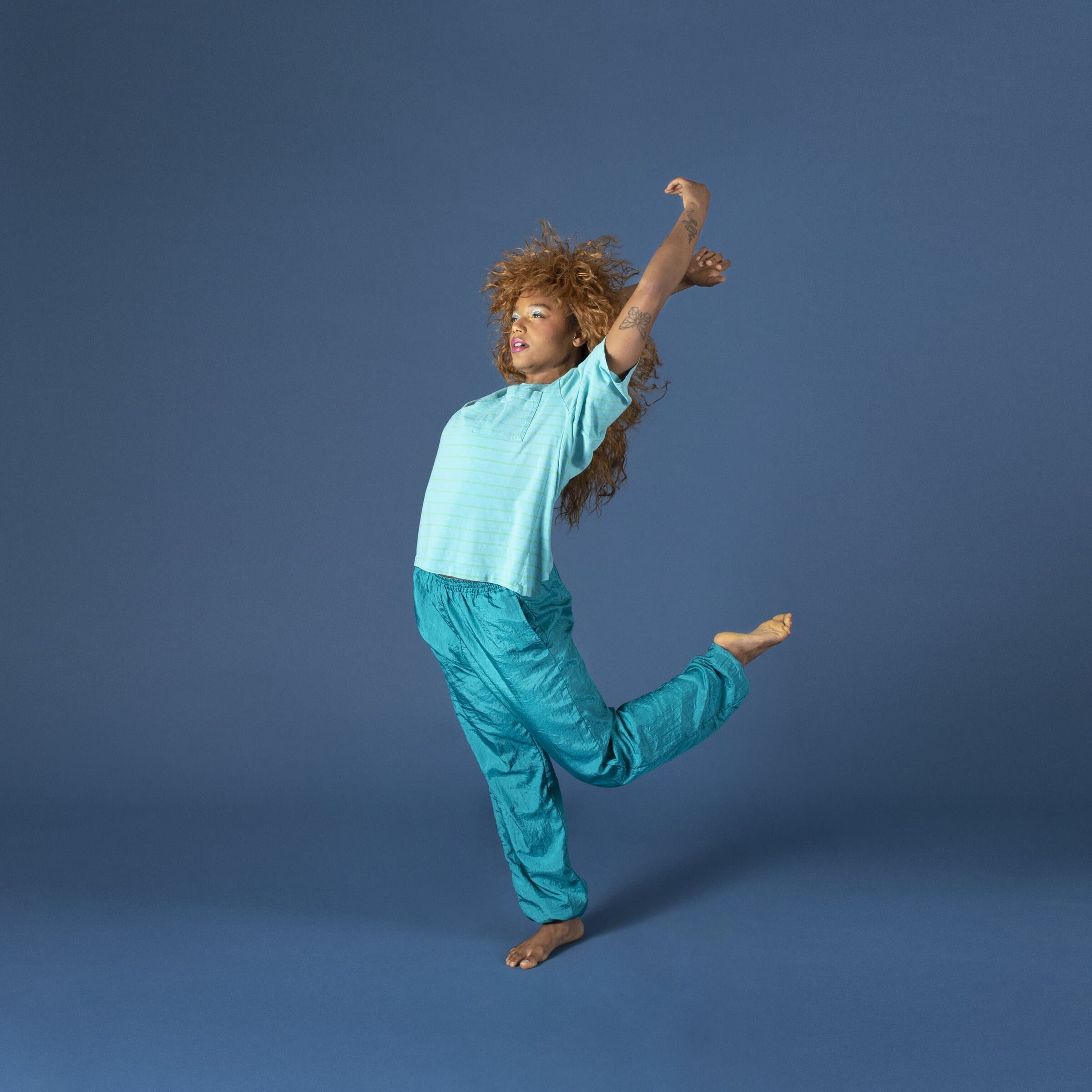 Dancer Gianna Theodore in a teal windbreaker outfit, leaping with arm raised against a blue backdrop in "Cassette Vol. 1."