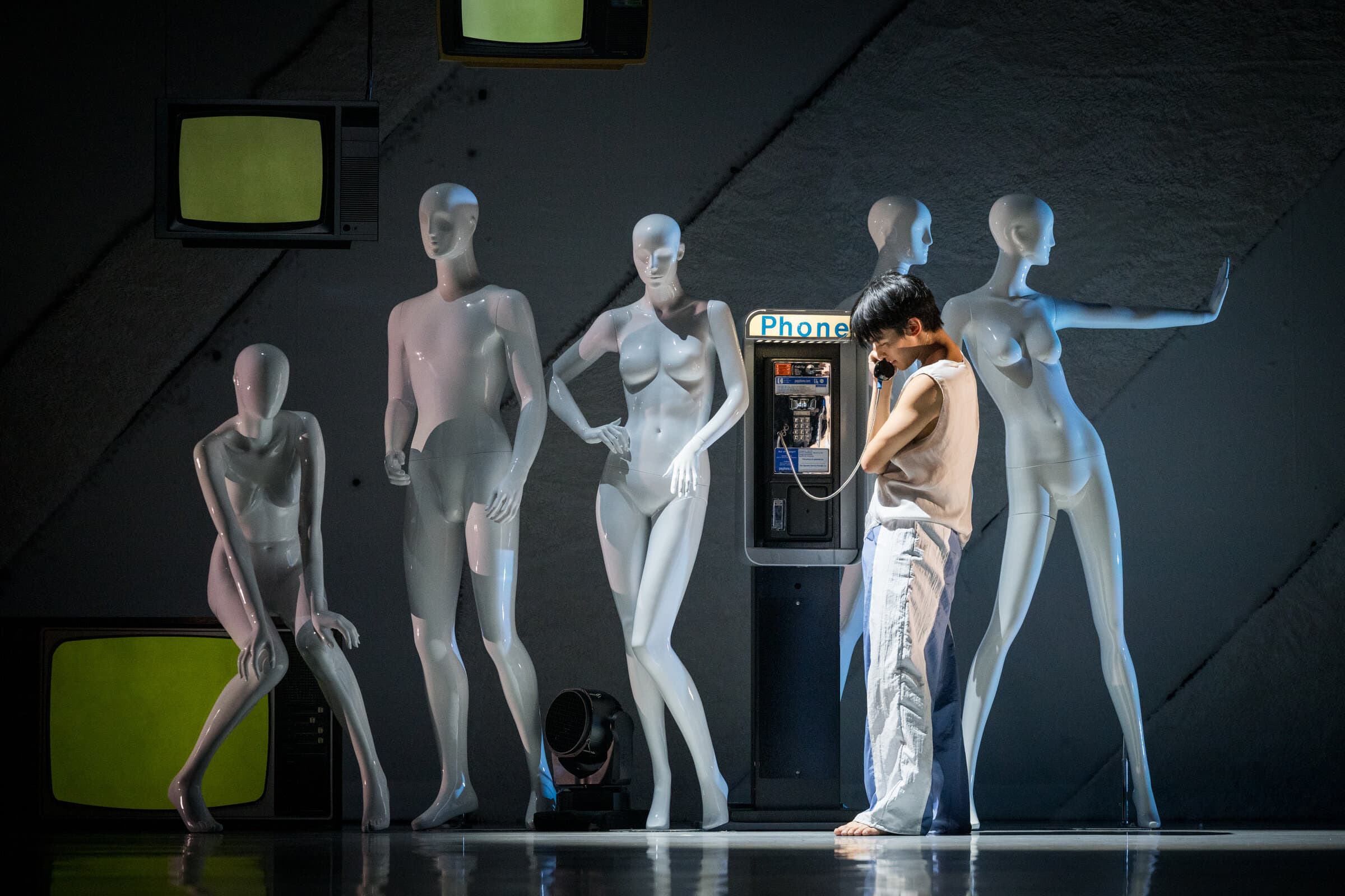 Dancer William Okajima on a payphone amid a row of glossy white mannequins and vintage TVs onstage.