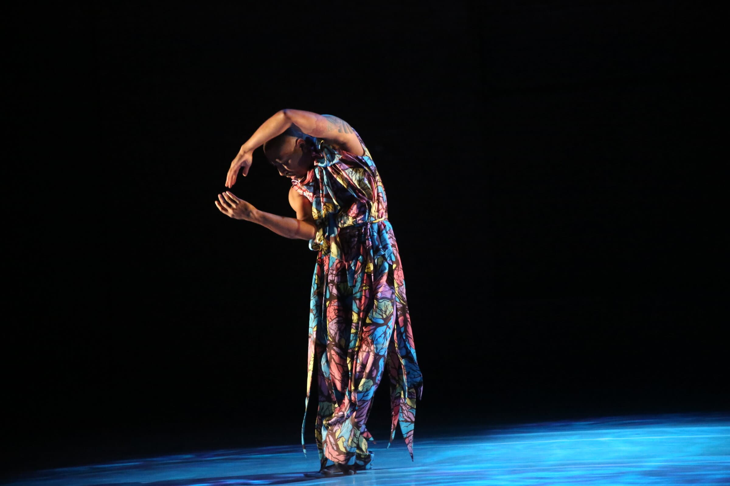 Dancer in a flowing, multicolor painted dress, bent forward with arms curved, lit in blue on a dark stage.