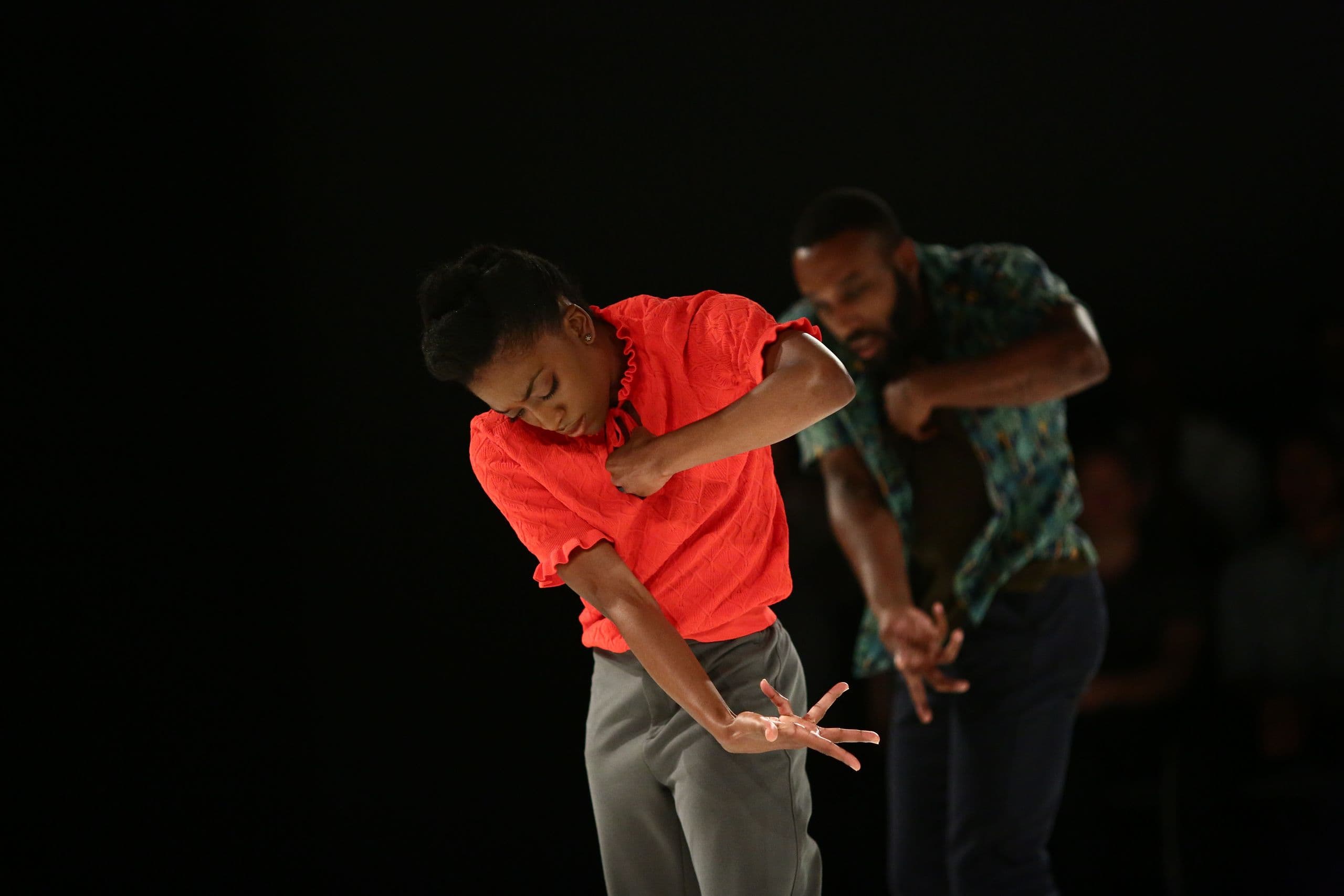 A dancer in a bright red top reaches downward mid-movement while a partner in a patterned shirt does the same behind her.