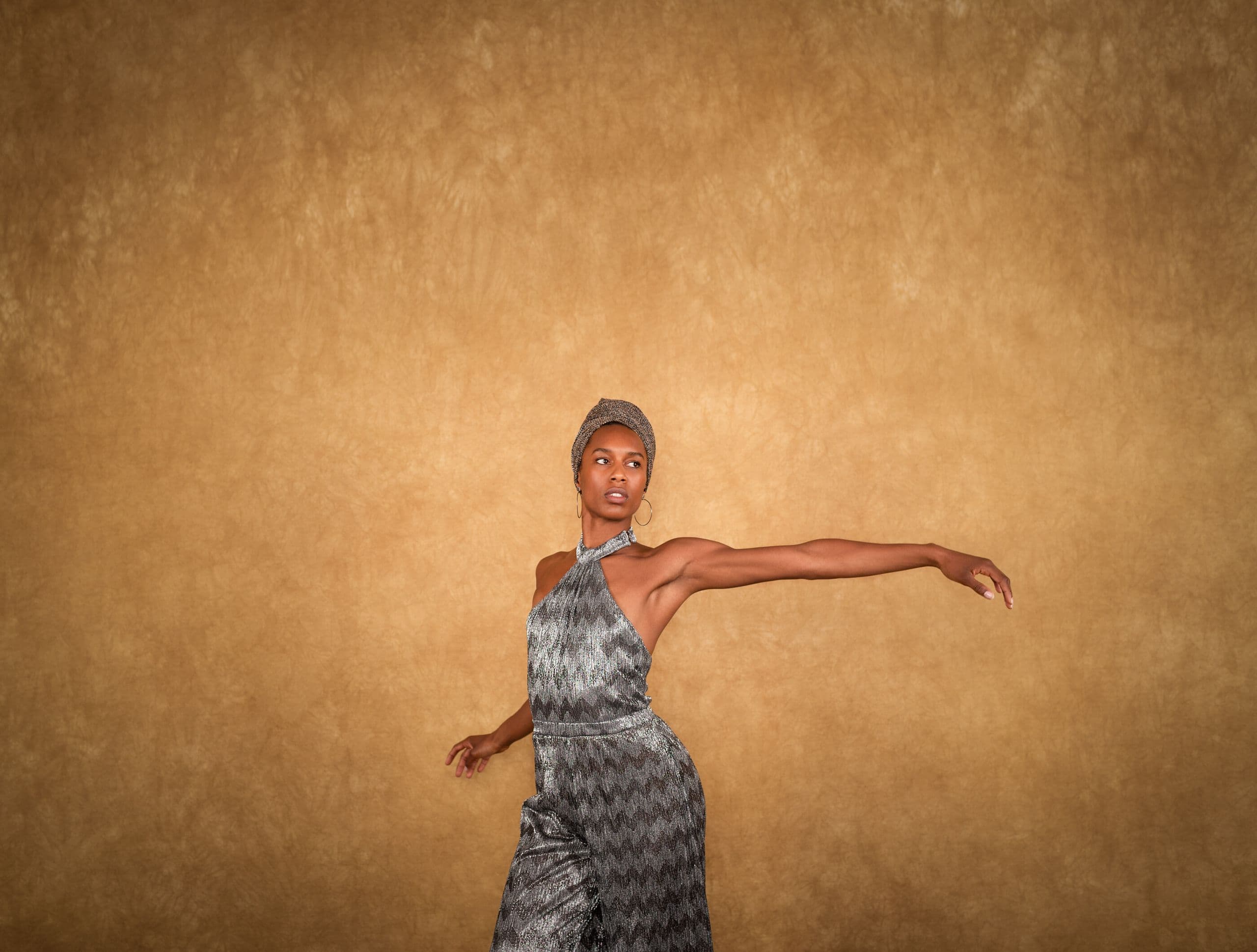 Catherine Kirk in a silver metallic jumpsuit and head wrap extends one arm outward against an amber backdrop