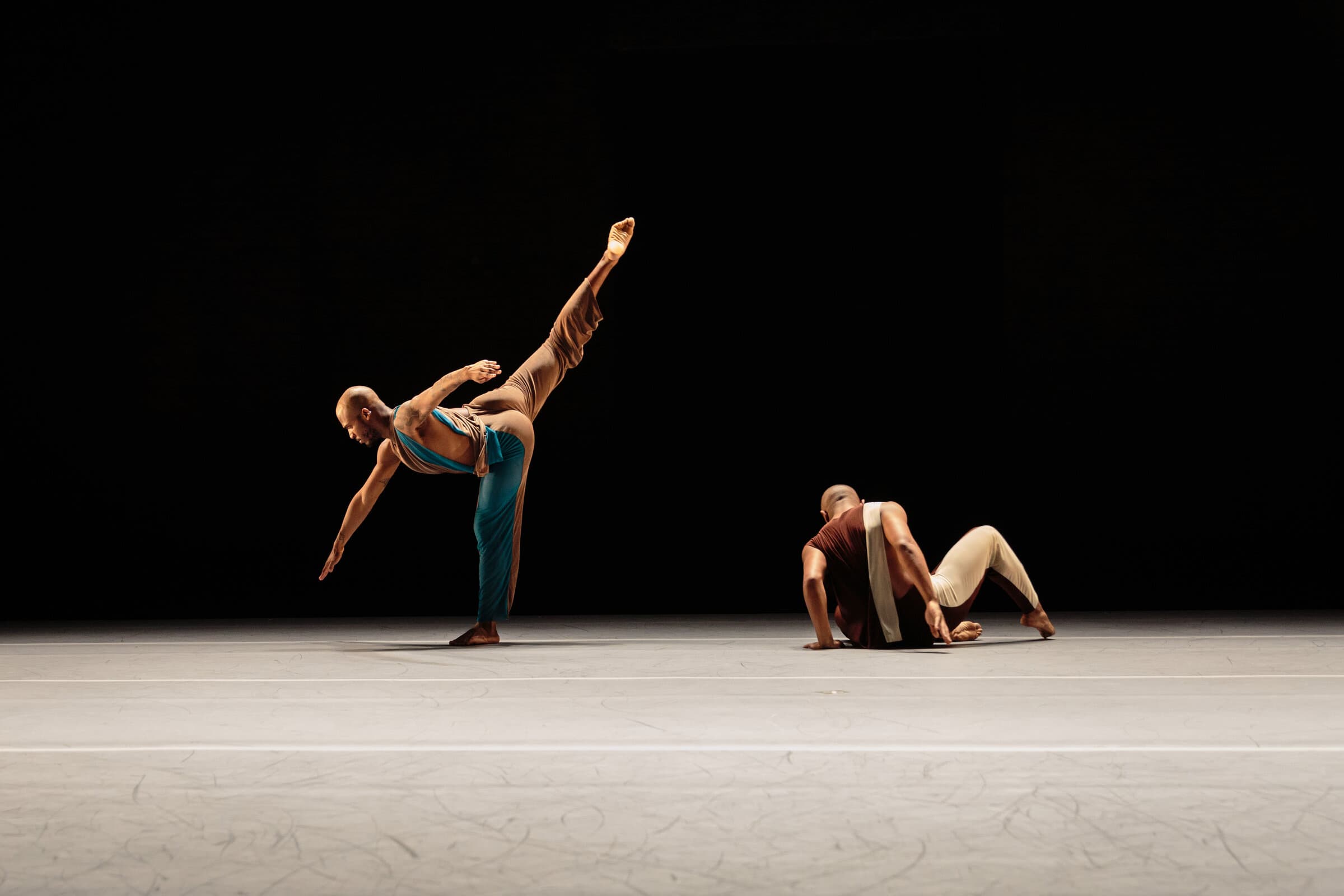 Dancers Donovan Reed and Jamaal Bowman in earth-toned costumes, one balanced in an extended arabesque, the other crouched on the floor.