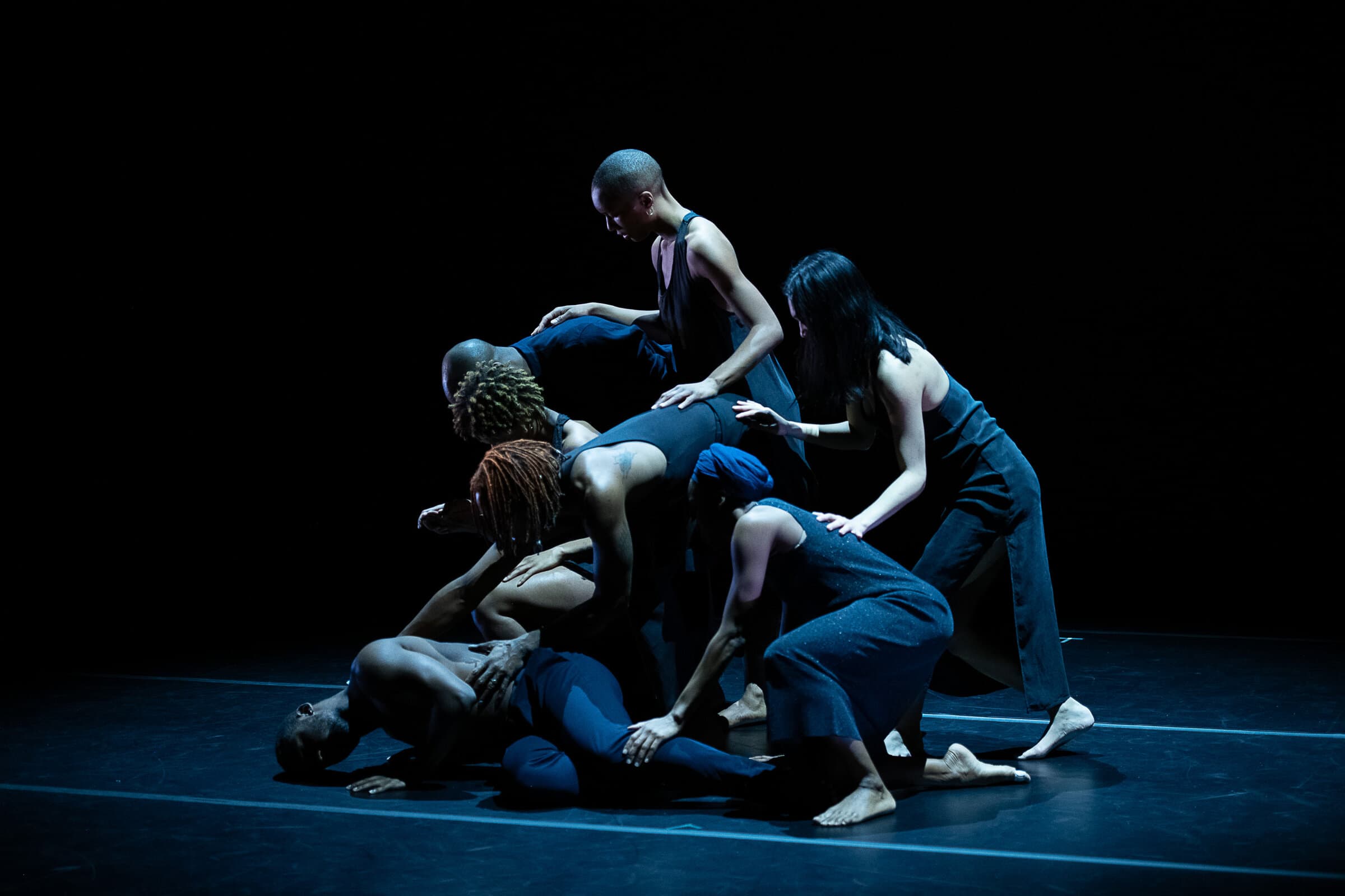 Group of dancers in dark blue costumes clustered together on a dim stage, supporting and reaching toward a dancer lying on the floor at the center.