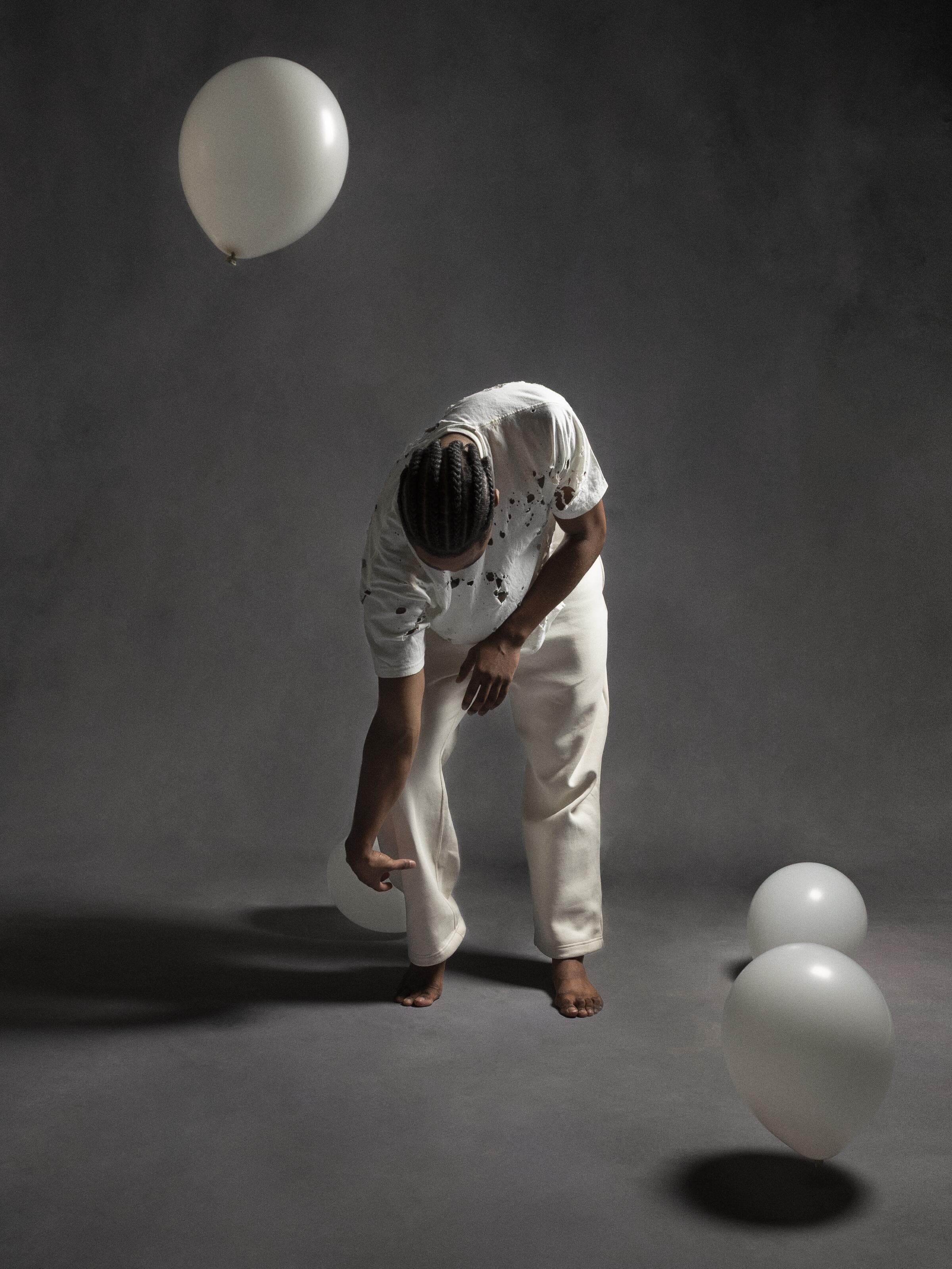 Dancer Destin Morisset bent forward in distressed white clothing, surrounded by floating white balloons in "White Space."