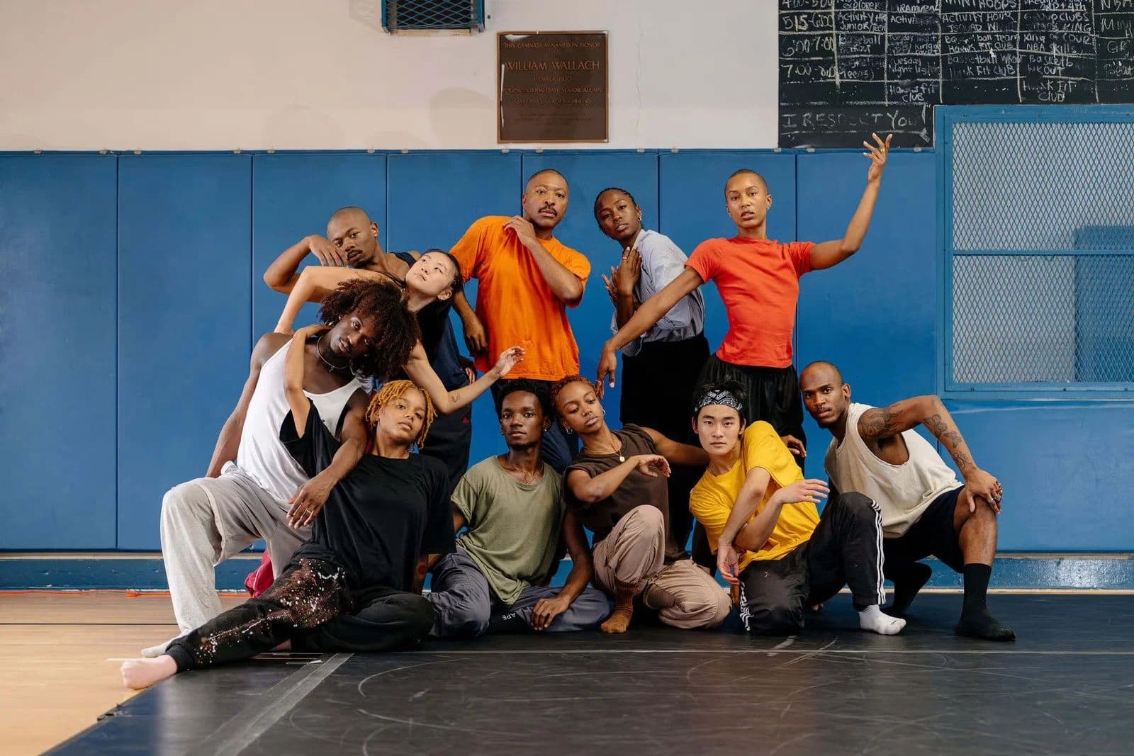 Group portrait of eleven dancers in colorful casual clothing, posed together in layered, expressive positions on a gym floor with blue padded walls and a chalkboard in the background.