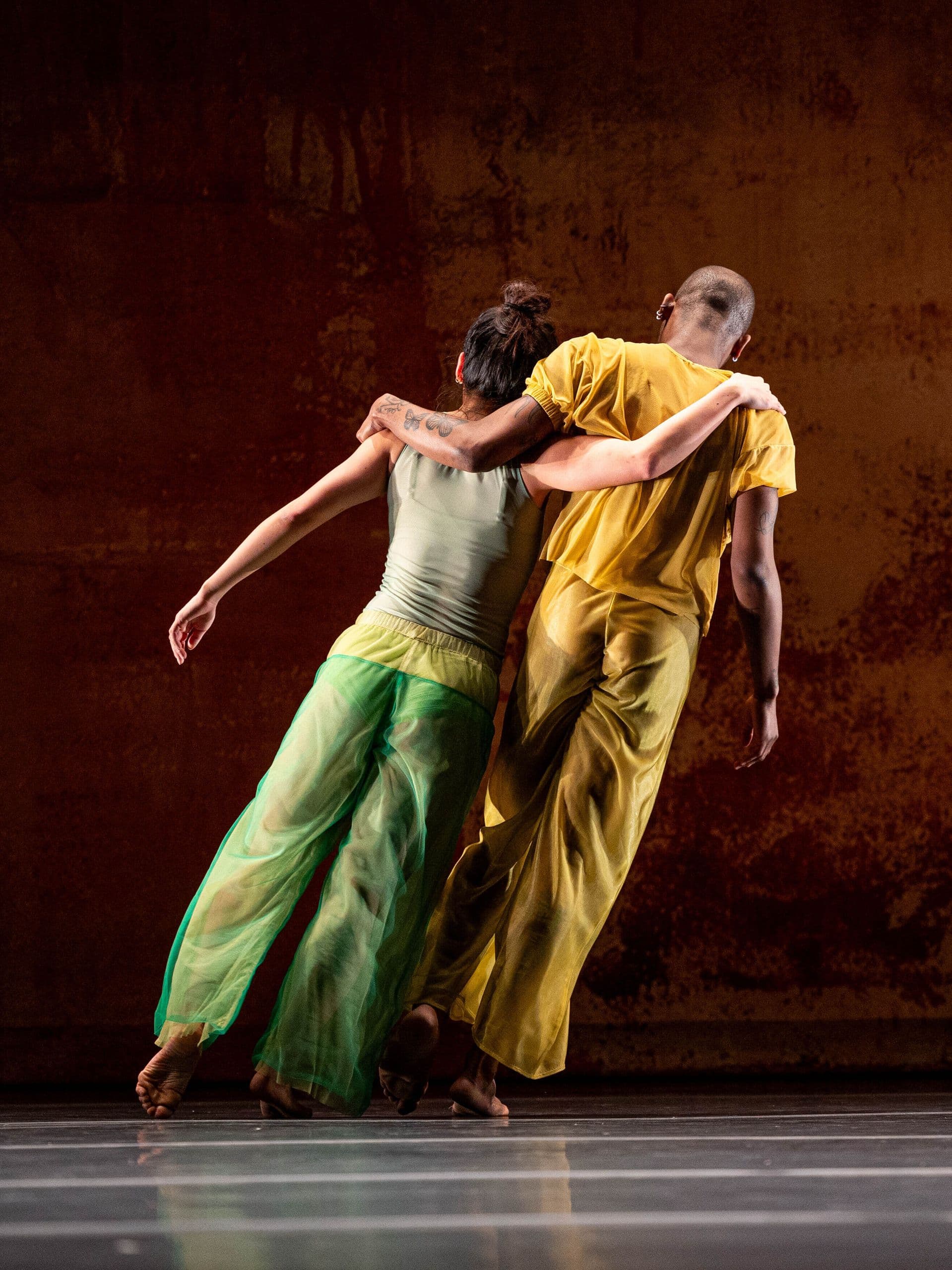 Two dancers seen from behind with arms around each other's shoulders, one in a green outfit and the other in yellow, standing on a stage with a weathered rust-colored backdrop