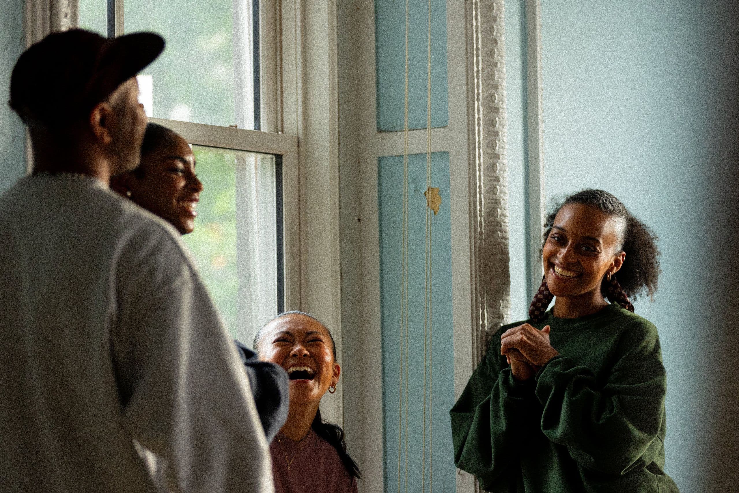 Four dancers laugh together in a sunlit studio with tall windows and pale blue walls, one in a green sweatshirt smiling toward the camera.