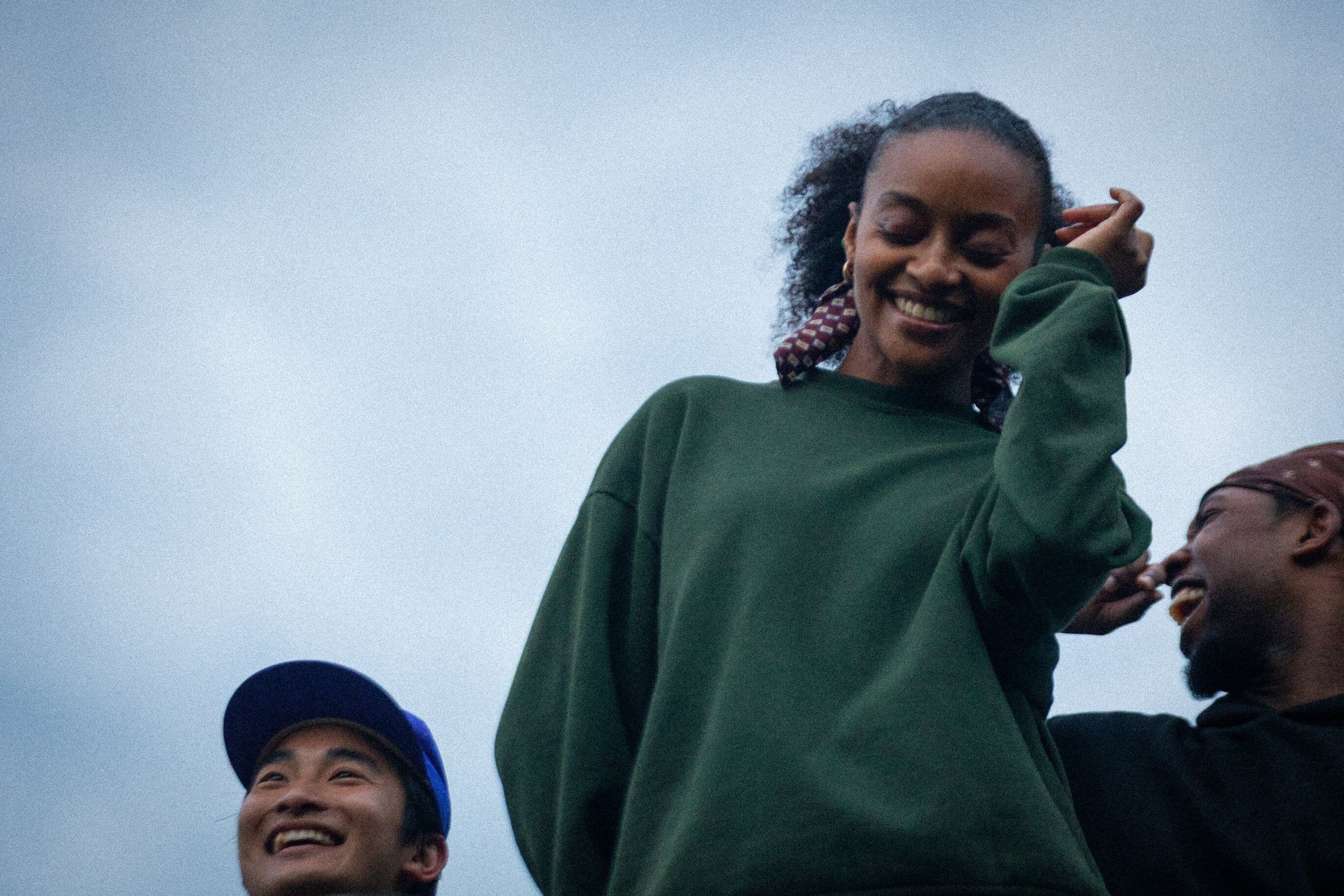 Alysia Johnson in an oversized green sweatshirt smiles with her eyes closed, touching her curly hair, with William Okajima in a blue cap and Destin Morissett in a red bandana visible beside her against a cloudy sky.