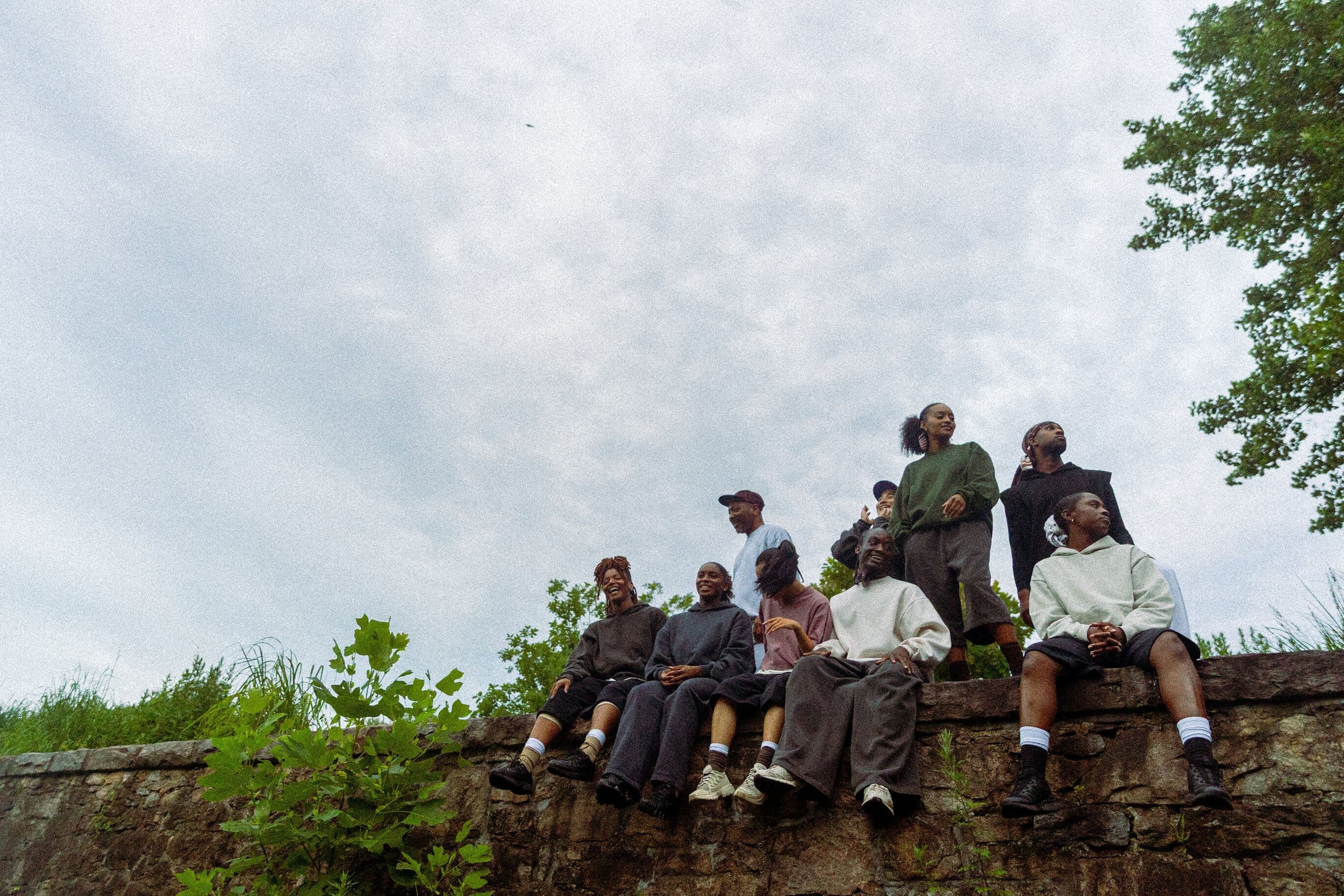 The company sitting and standing on a stone wall, looking up at an overcast sky, surrounded by green trees and grass, wearing casual oversized streetwear in earth tones