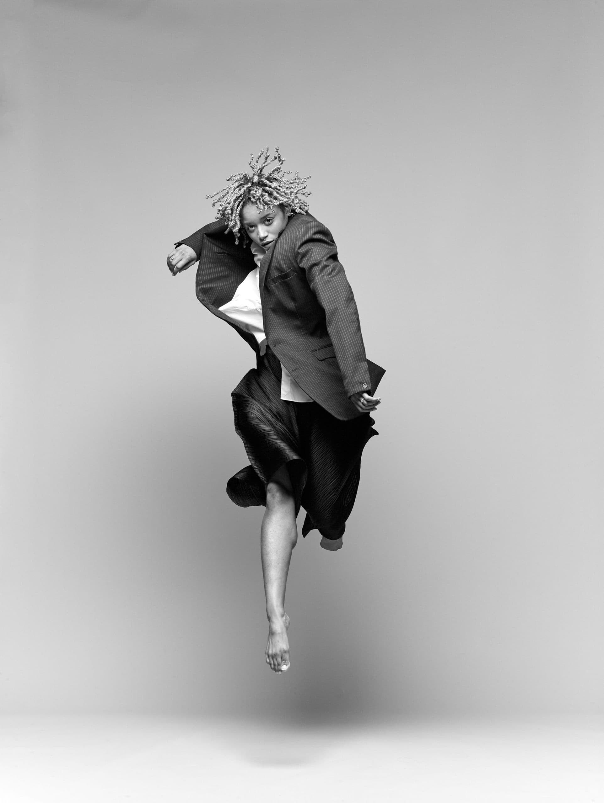 Black-and-white photo of a dancer captured mid-jump in a pinstriped blazer and pleated skirt, hair flying upward, gazing directly at the camera against a plain grey background.