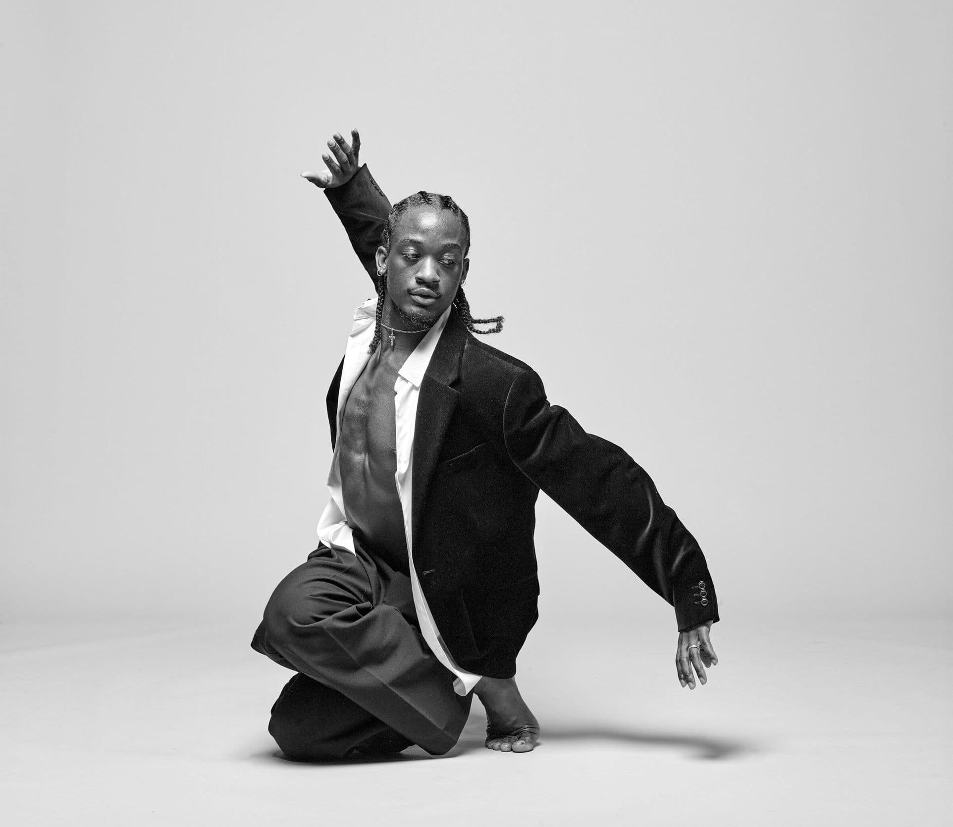 Black-and-white photo of a dancer kneeling on one knee in a black blazer and white open shirt, mid-motion with one arm raised and fingers splayed, against a plain grey background