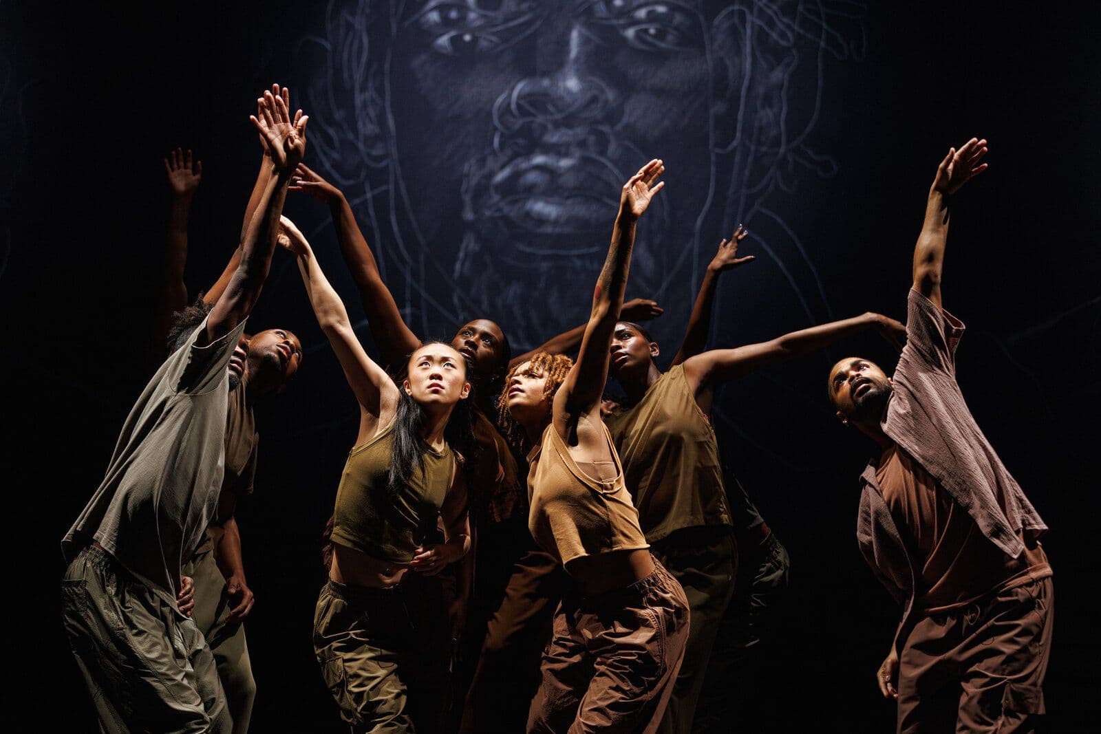 A group of dancers in earth-toned clothing reach their arms upward together, gazing skyward, silhouetted against a large illustrated portrait of a face projected on the backdrop behind them.