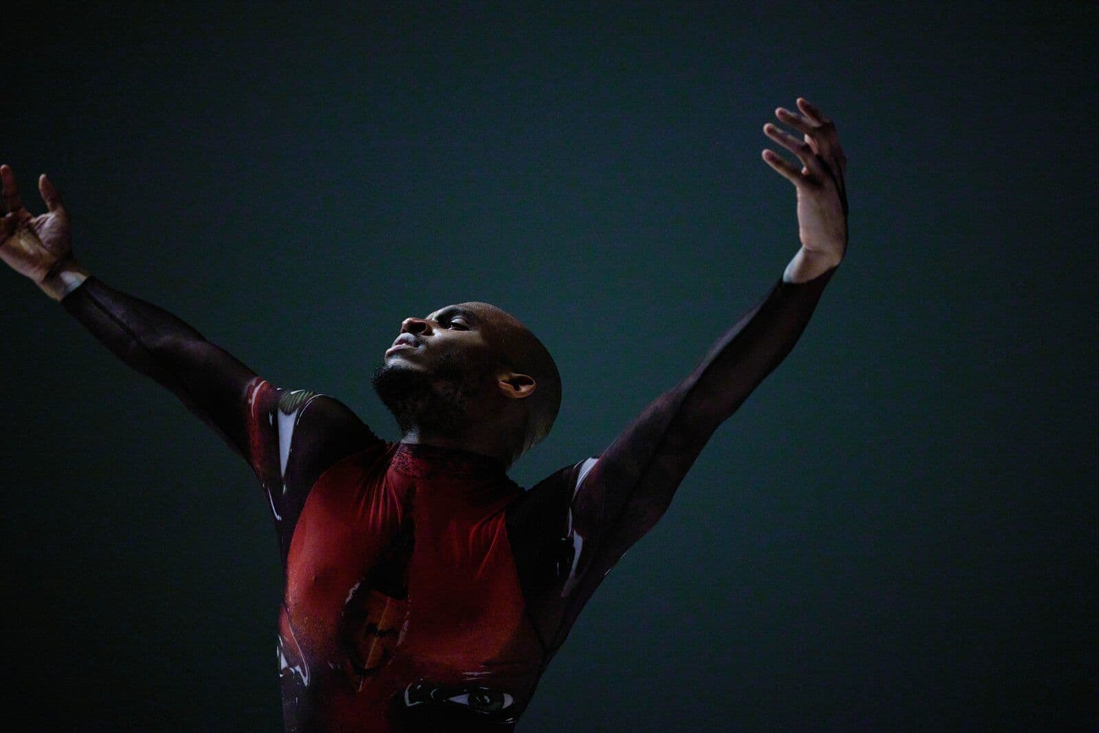 Dancer reaches both arms upward in a graphic red-and-black patterned costume, lit dramatically against a dark teal background.