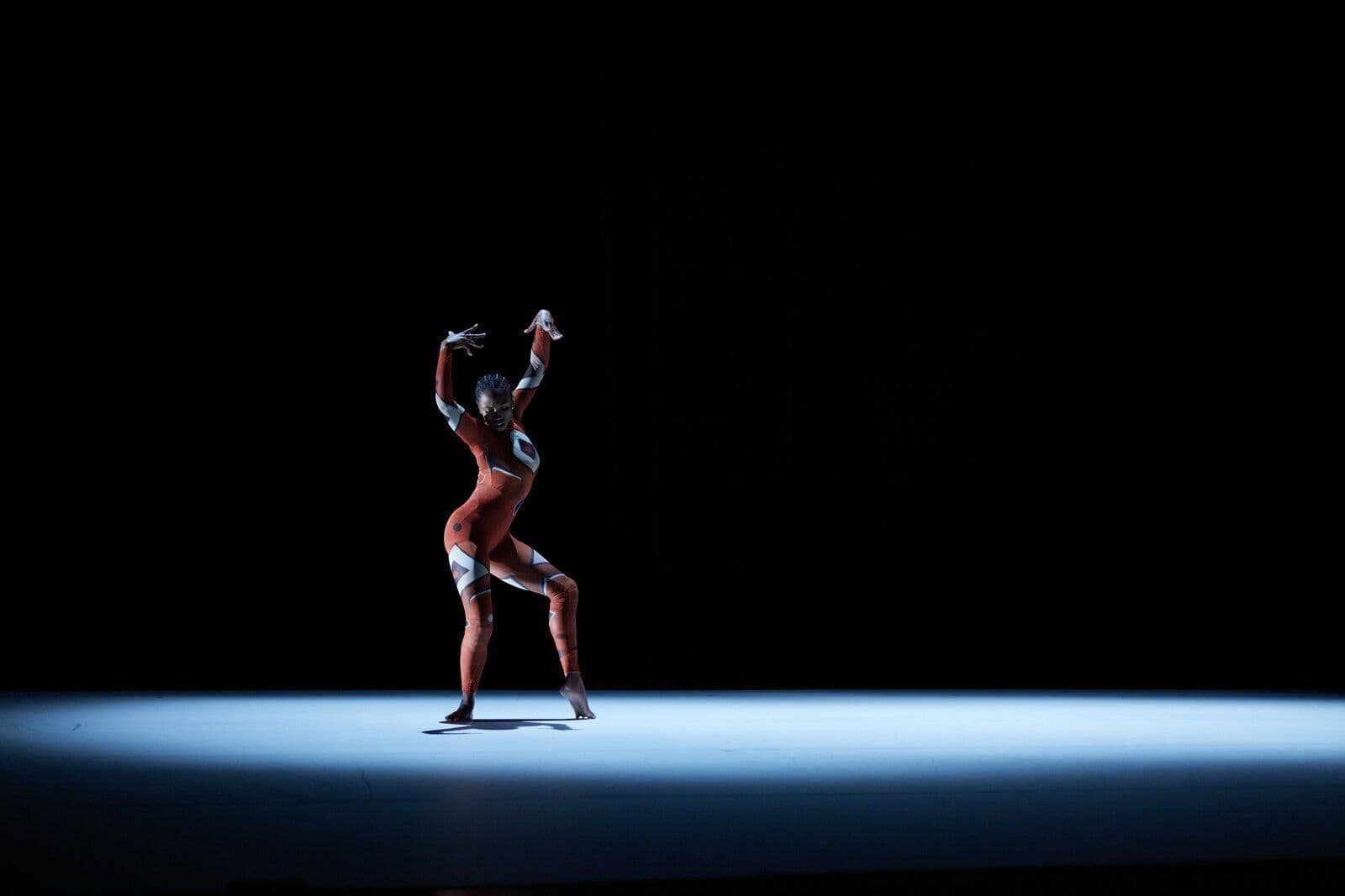 A dancer performs a grounded, angular pose in a black-and-red patterned costume, illuminated by a single spotlight against a dark stage.