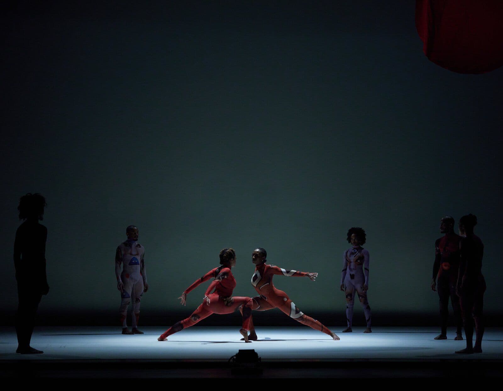 Two dancers in red patterned bodysuits face off in a low lunge under a spotlight, surrounded by silhouetted ensemble members in patterned costumes on a dark stage.